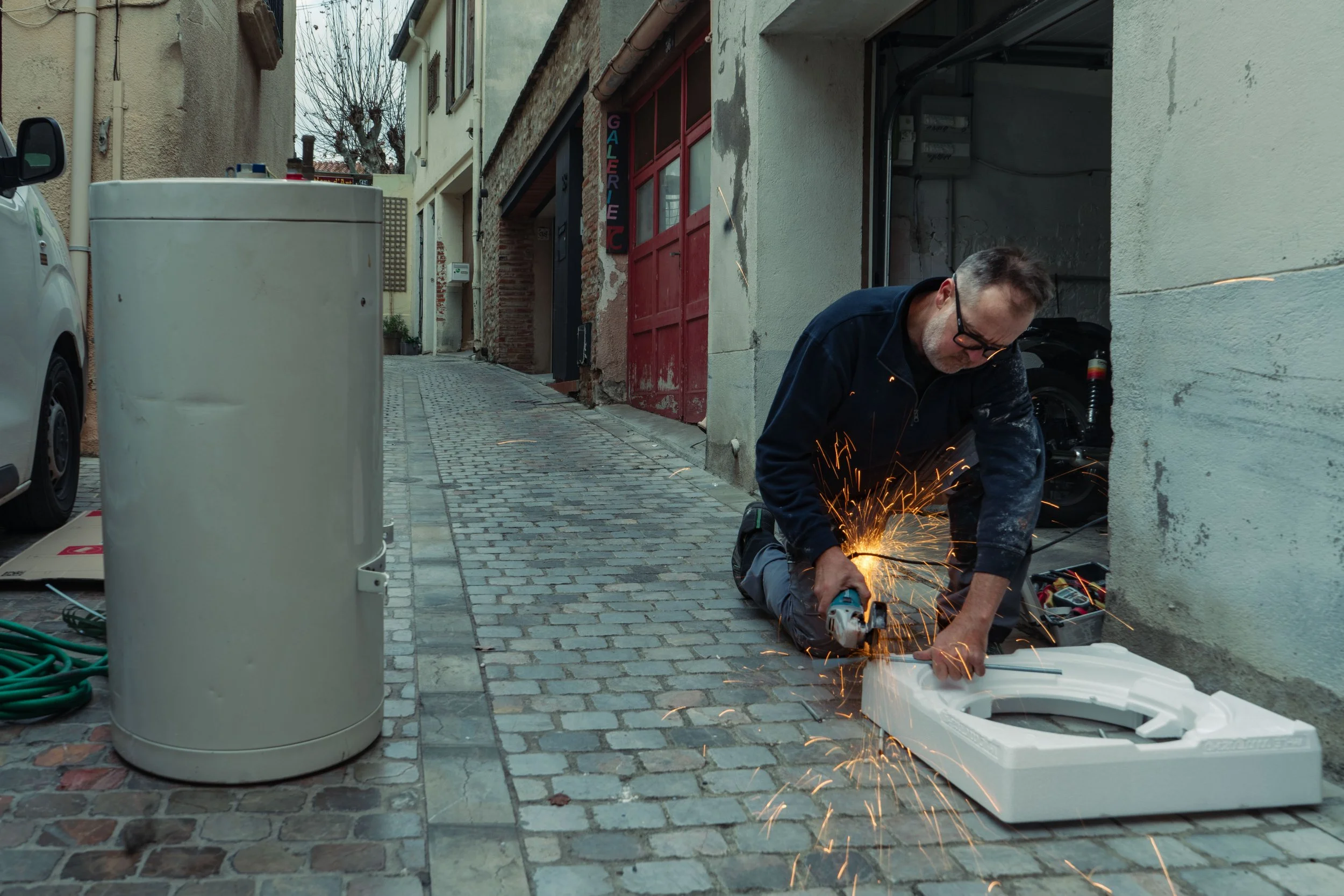 Un homme en train de découper un objet en utilisant une meuleuse d'angle, avec des étincelles volant autour. Il est agenouillé dans une ruelle pavée, à côté d'un cylindre blanc et devant un bâtiment. Derrière lui, il y a une camionnette garée.