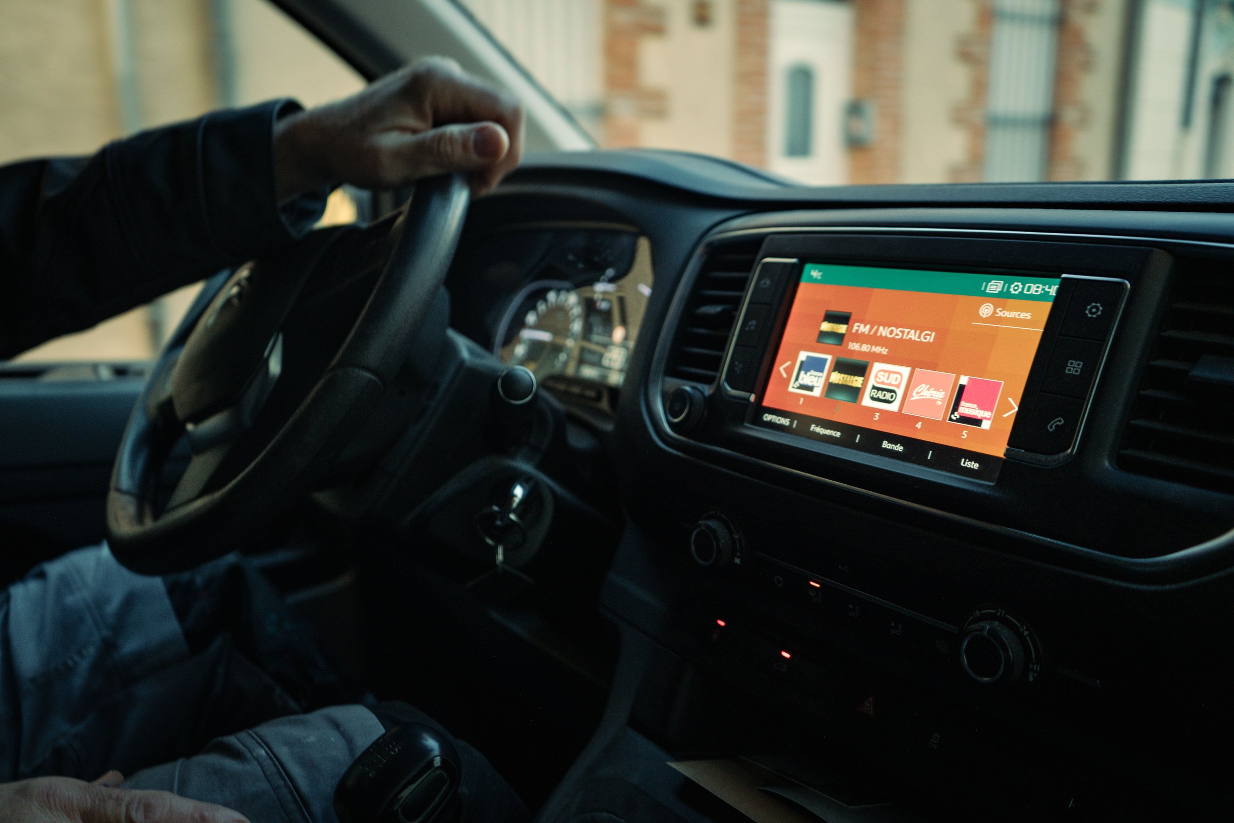 Intérieur d'une voiture avec un écran affichant les stations de radio. Une personne tient le volant.