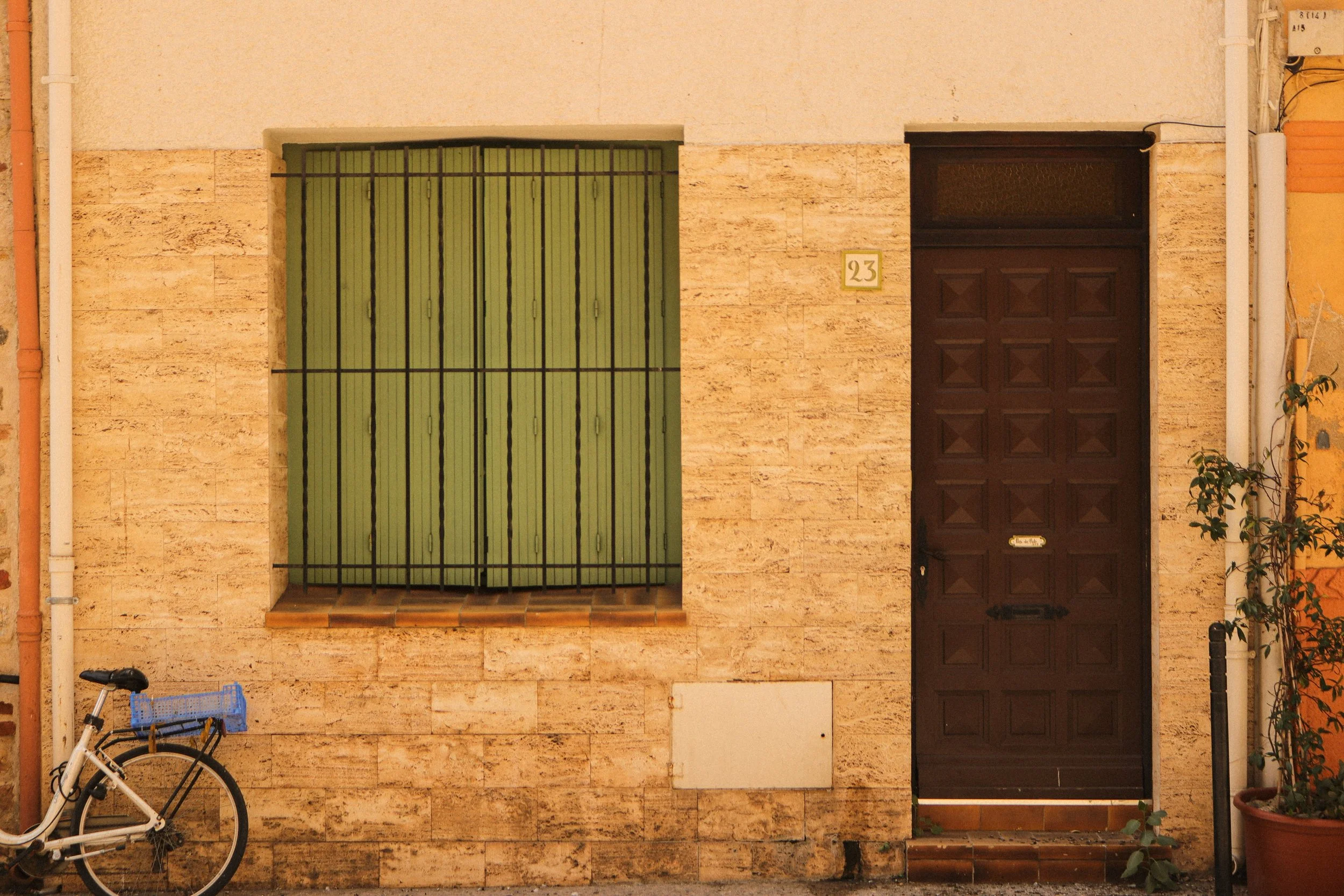 Façade de maison avec porte en bois, fenêtre avec volets verts et grille en métal, vélo garé devant.