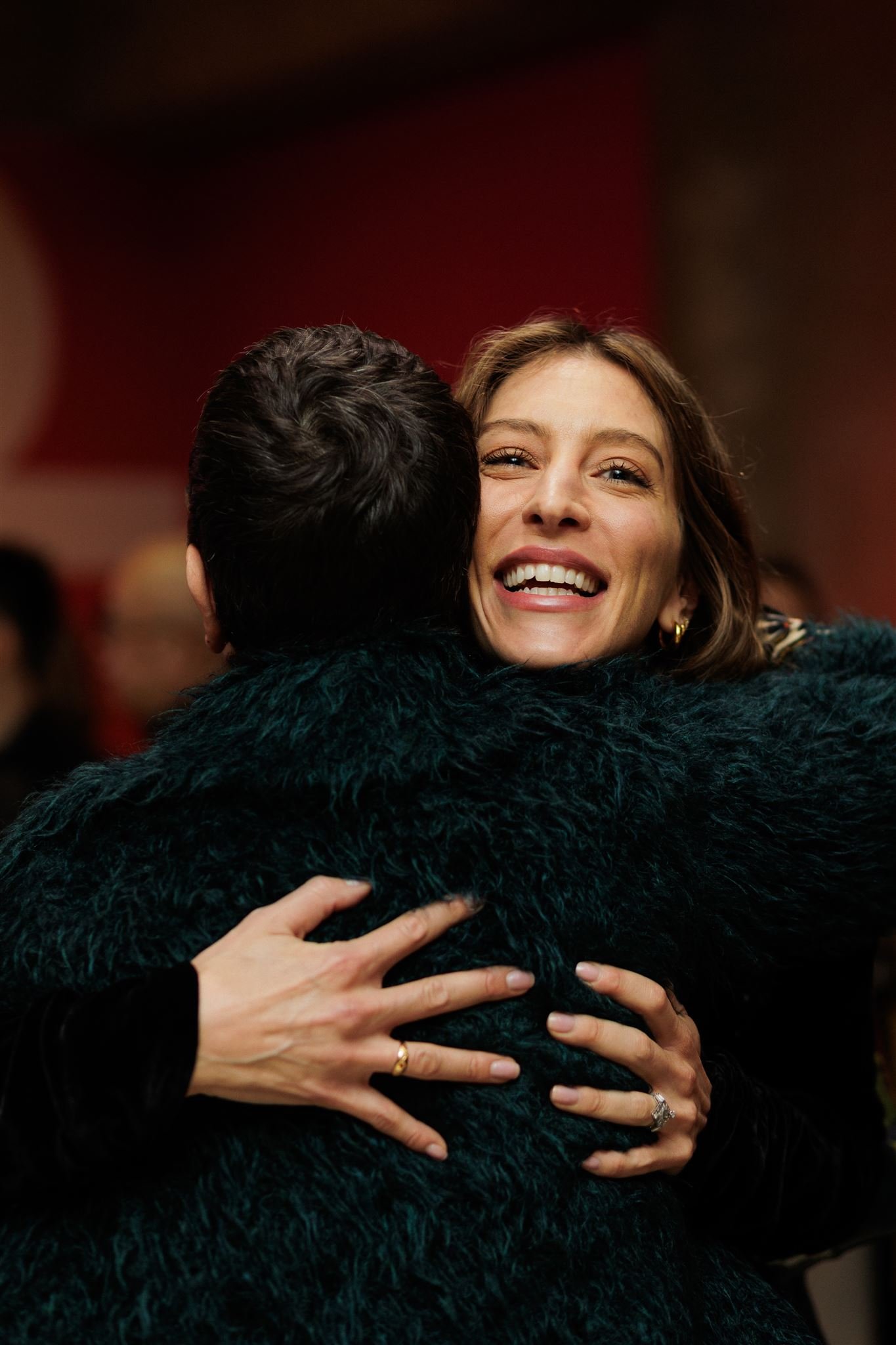 A woman in a green faux fur coat hugging a laughing friend at a London art gallery opening event.