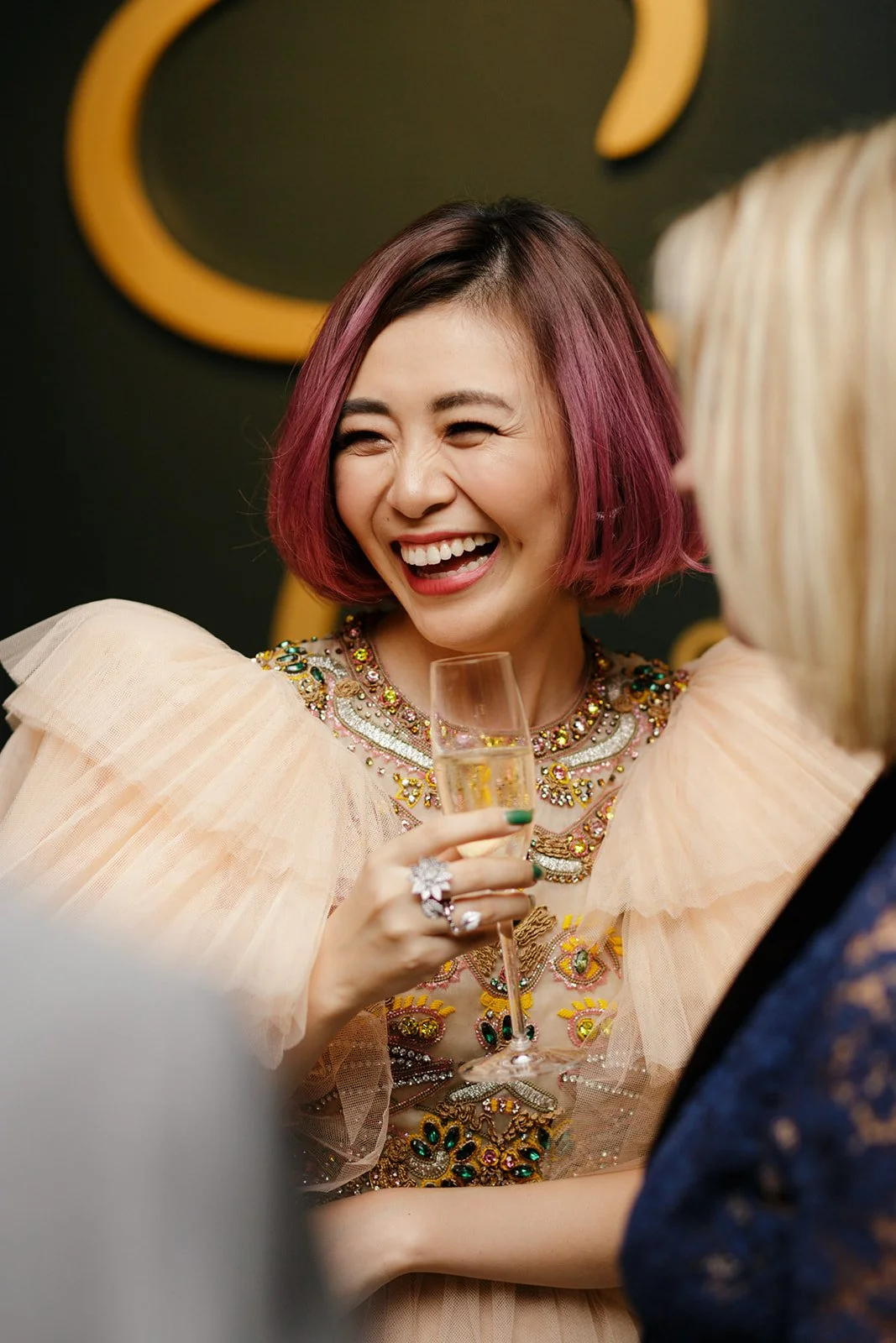 A woman with pink hair laughing while holding a champagne glass at a high-end fashion event.