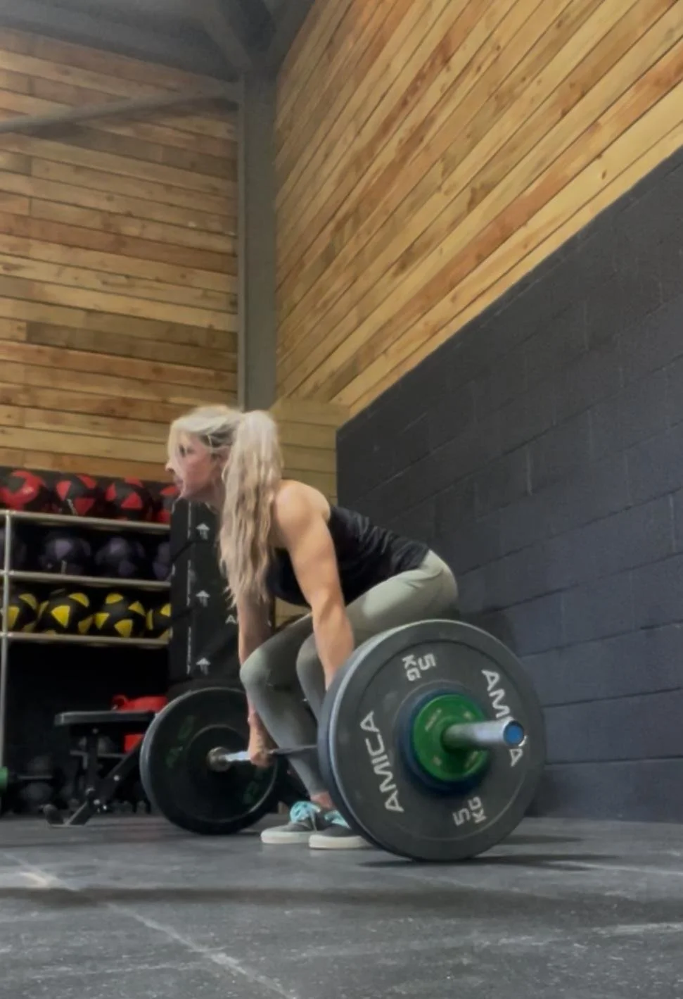 A woman with blonde hair lifting a barbell loaded with weight plates in a gym with wooden walls and a black painted wall. Heather at HPT Personal training