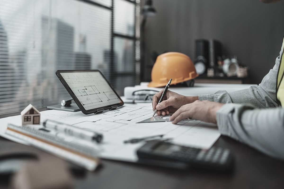 An architect working on blueprints at a desk, with a computer tablet displaying a floor plan, a yellow safety helmet, and a small model house.