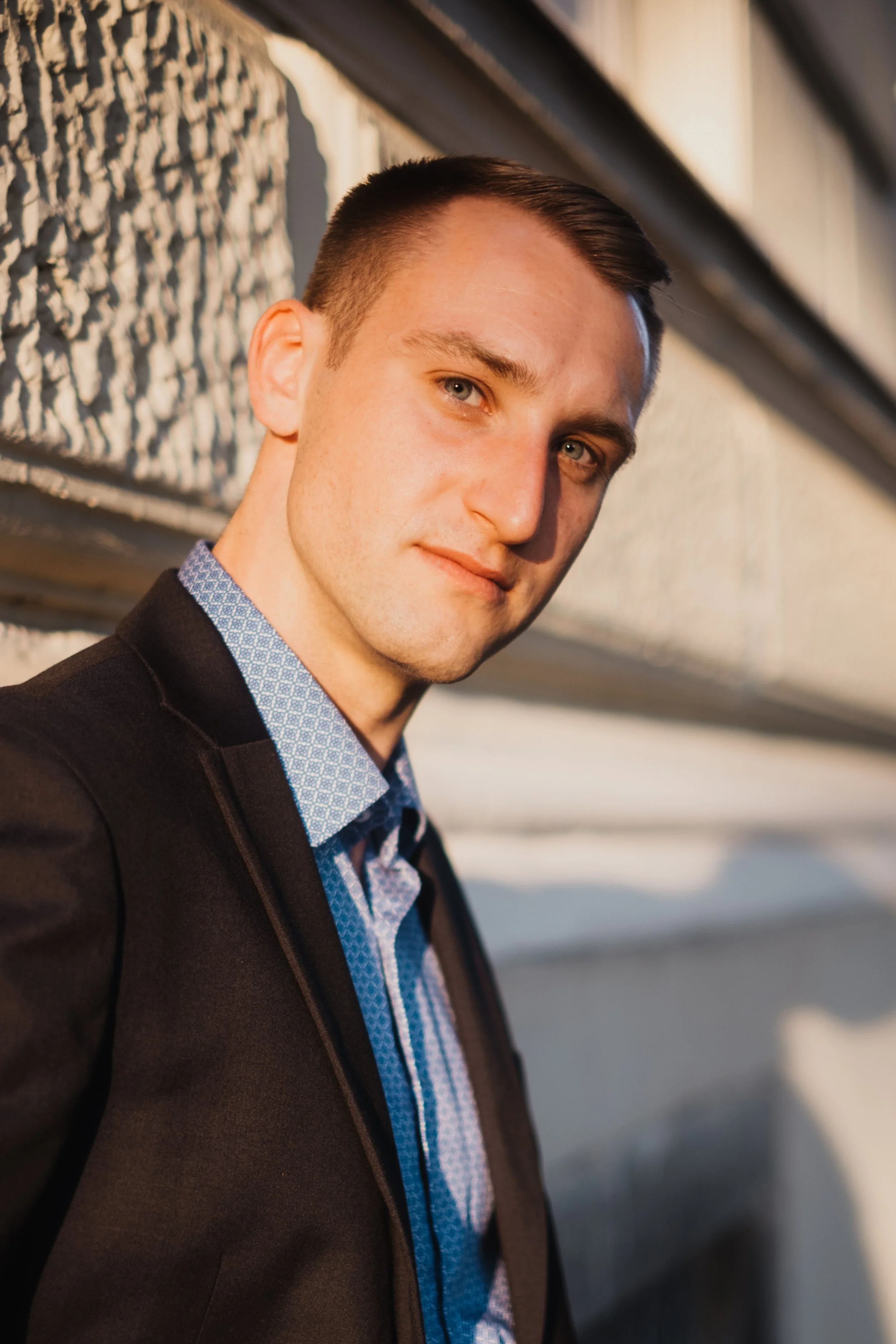 Junger Mann in dunklem Anzug und blauer Hemd lehnend an einer grauen Wand, Blick in die Kamera, Sonnenlicht auf seinem Gesicht, urbaner Hintergrund.