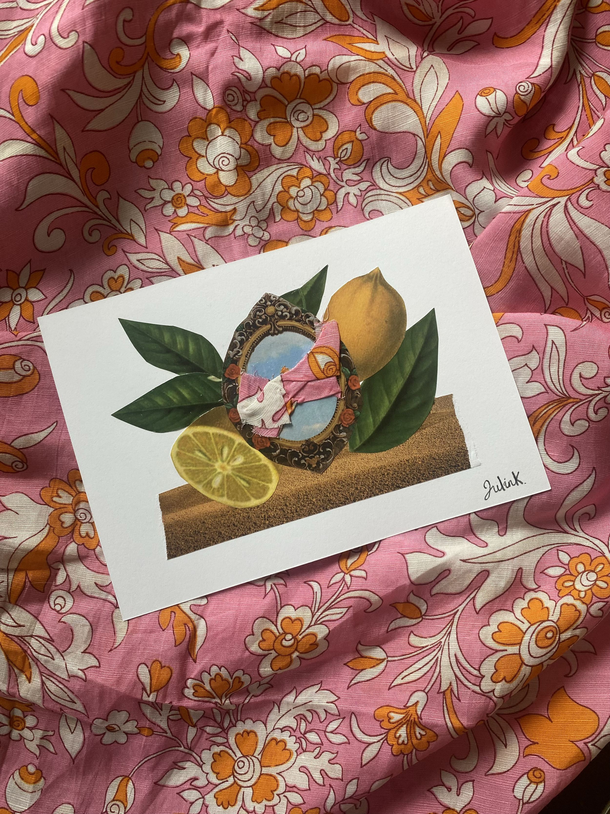 Photo d'une illustration collage représentant un cadre décoré de fleurs et de fruits, avec un fond de terre ou sable, sur un tissu rose avec un motif floral orange et blanc. Collages contemporains surréalistes par Julia Chameroy