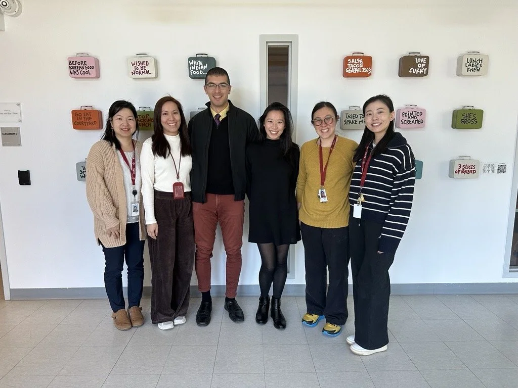 Six people, professionally dressed, stand together smiling in front of a white wall featuring an artistic display of lunch boxes.