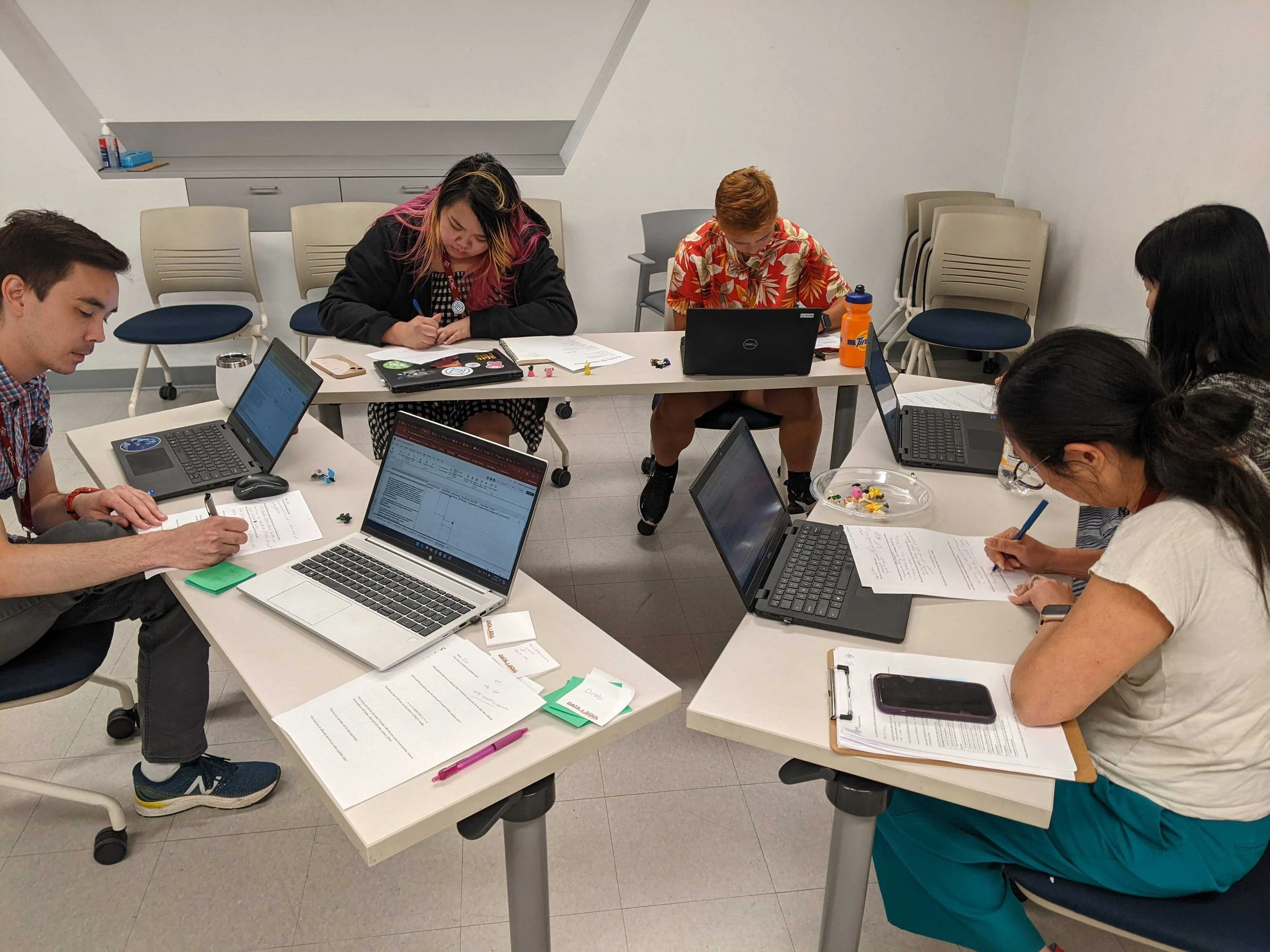 Five people sitting around a table, working on paper with laptops nearby.
