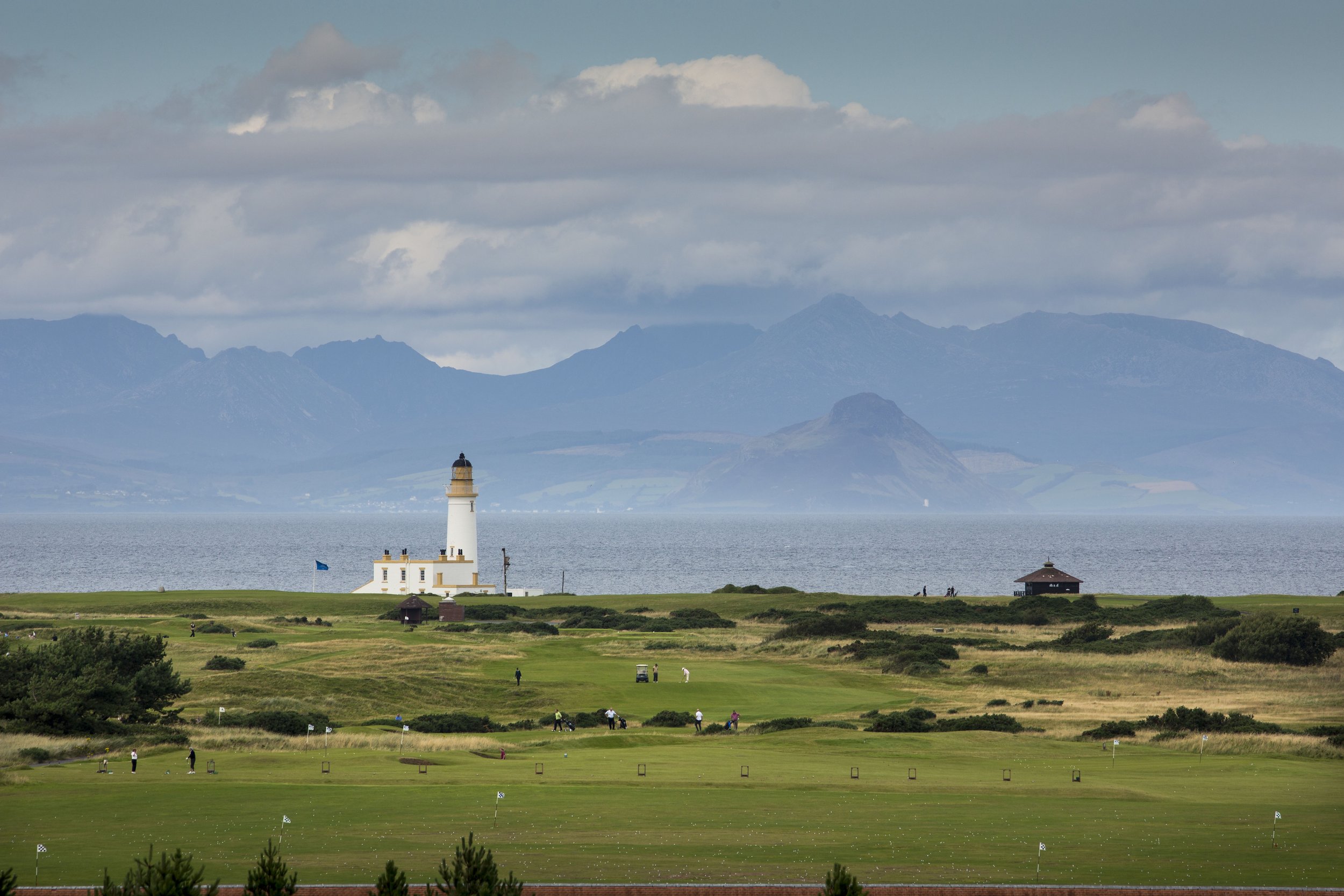 Coastal landscape with golf course and lighthouse, distant mountains and cloudy sky.