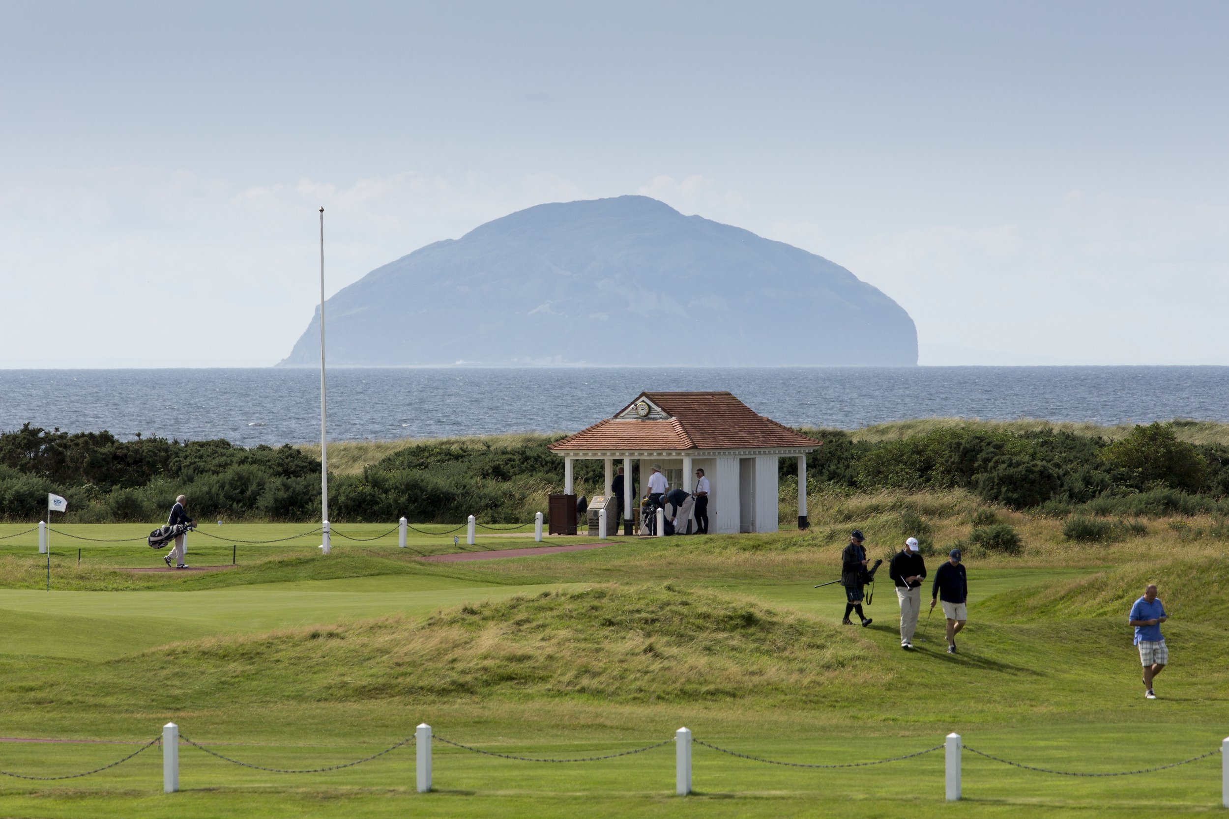 Golfers on a golf course with a view of the ocean and an island in the distance. A small white pavilion with a red roof is in the foreground.