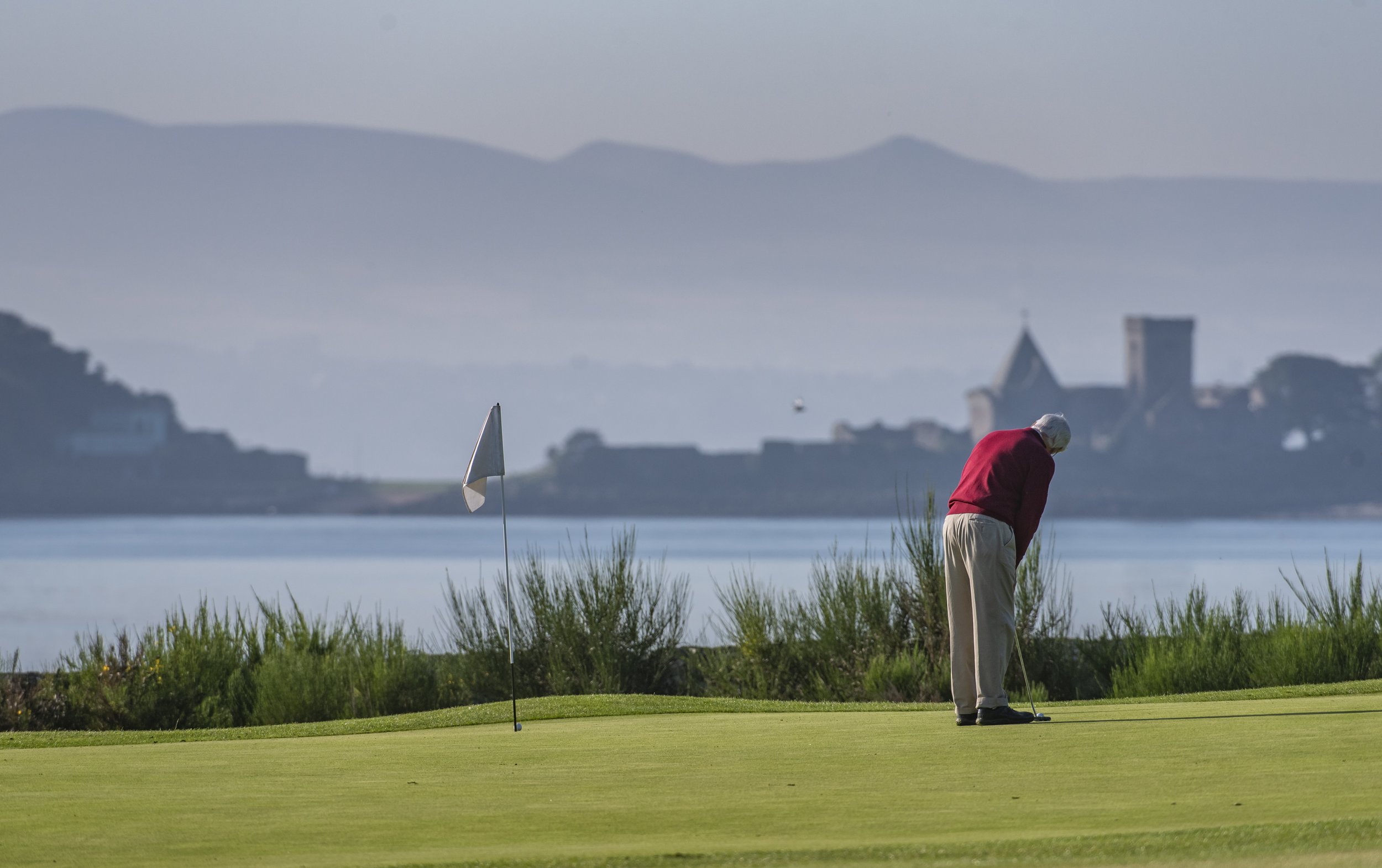 Golfer putting on a green near a lake with castle in background.