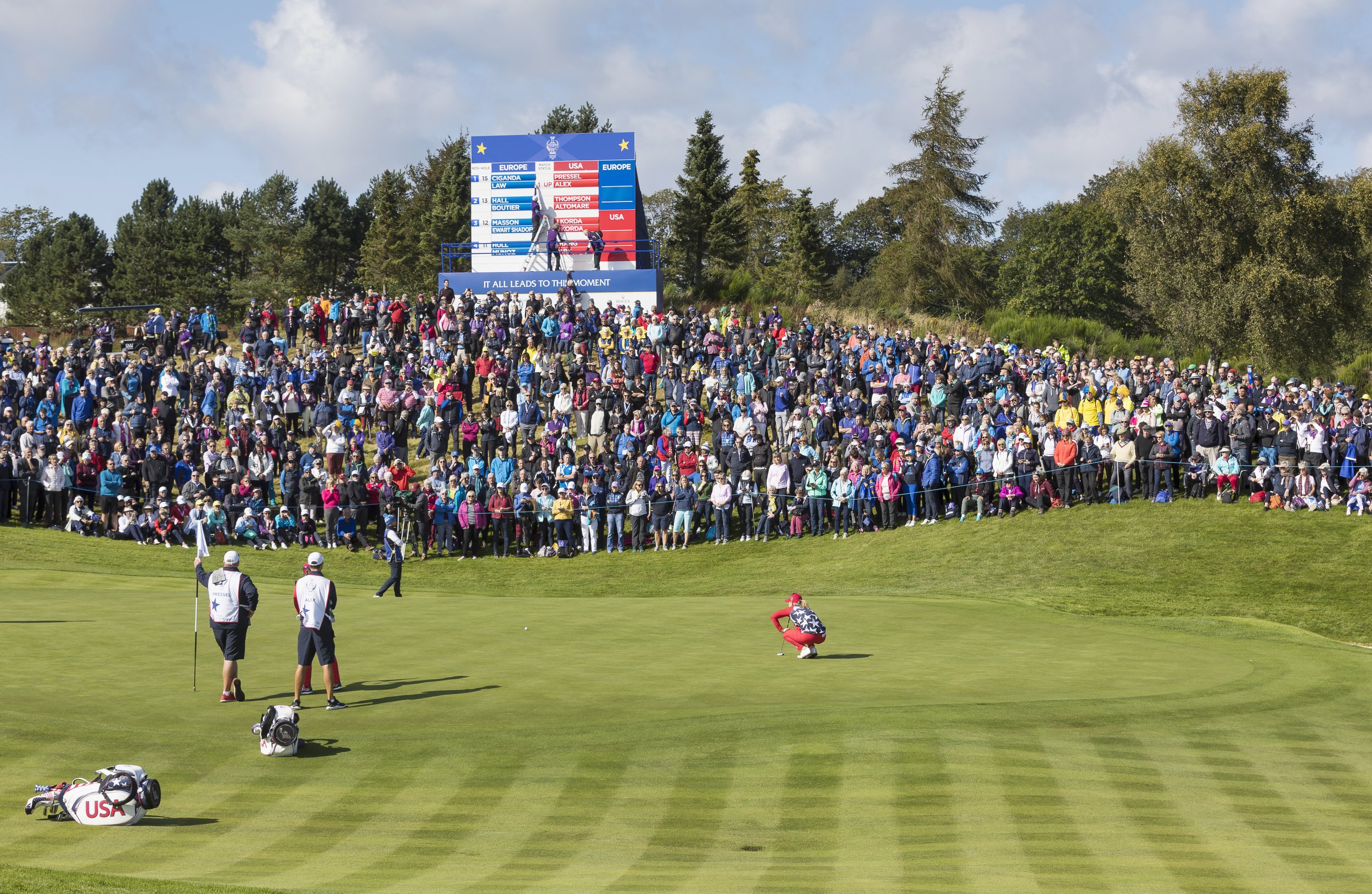 A golfer on a green surrounded by a large crowd, with two caddies standing nearby and a leaderboard in the background.