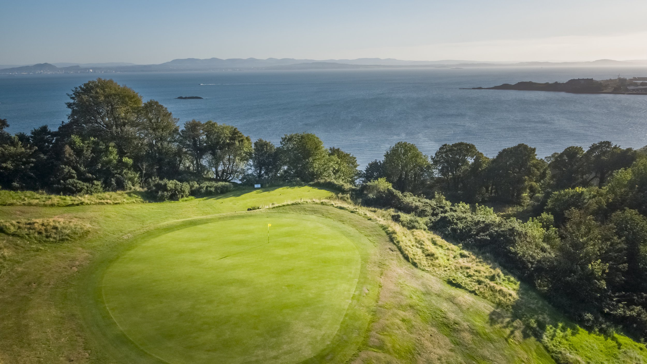 Scenic view of a golf course with a green overlooking a body of water, surrounded by trees and an expansive landscape.