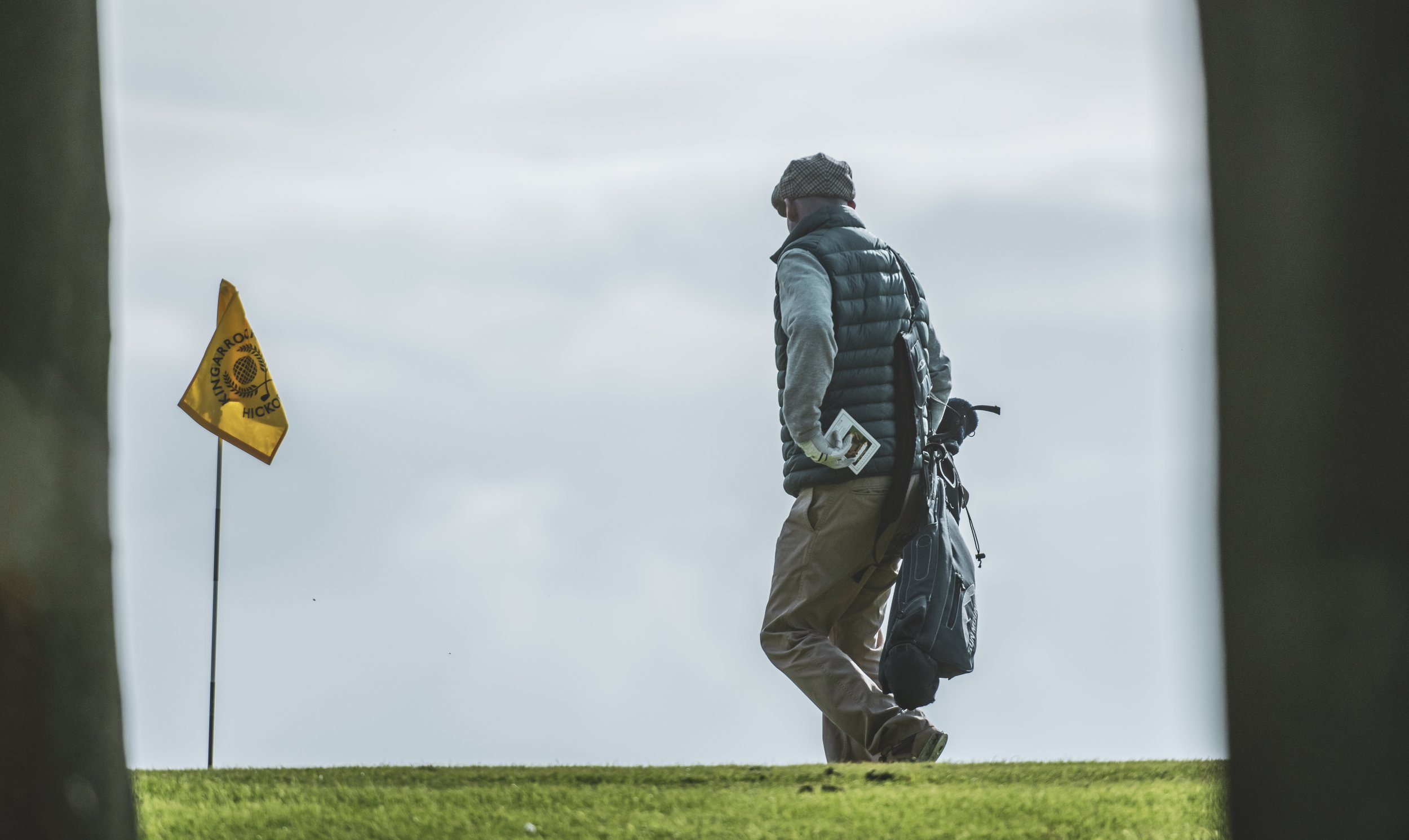 Golfer carrying clubs near a yellow flag on green grass.