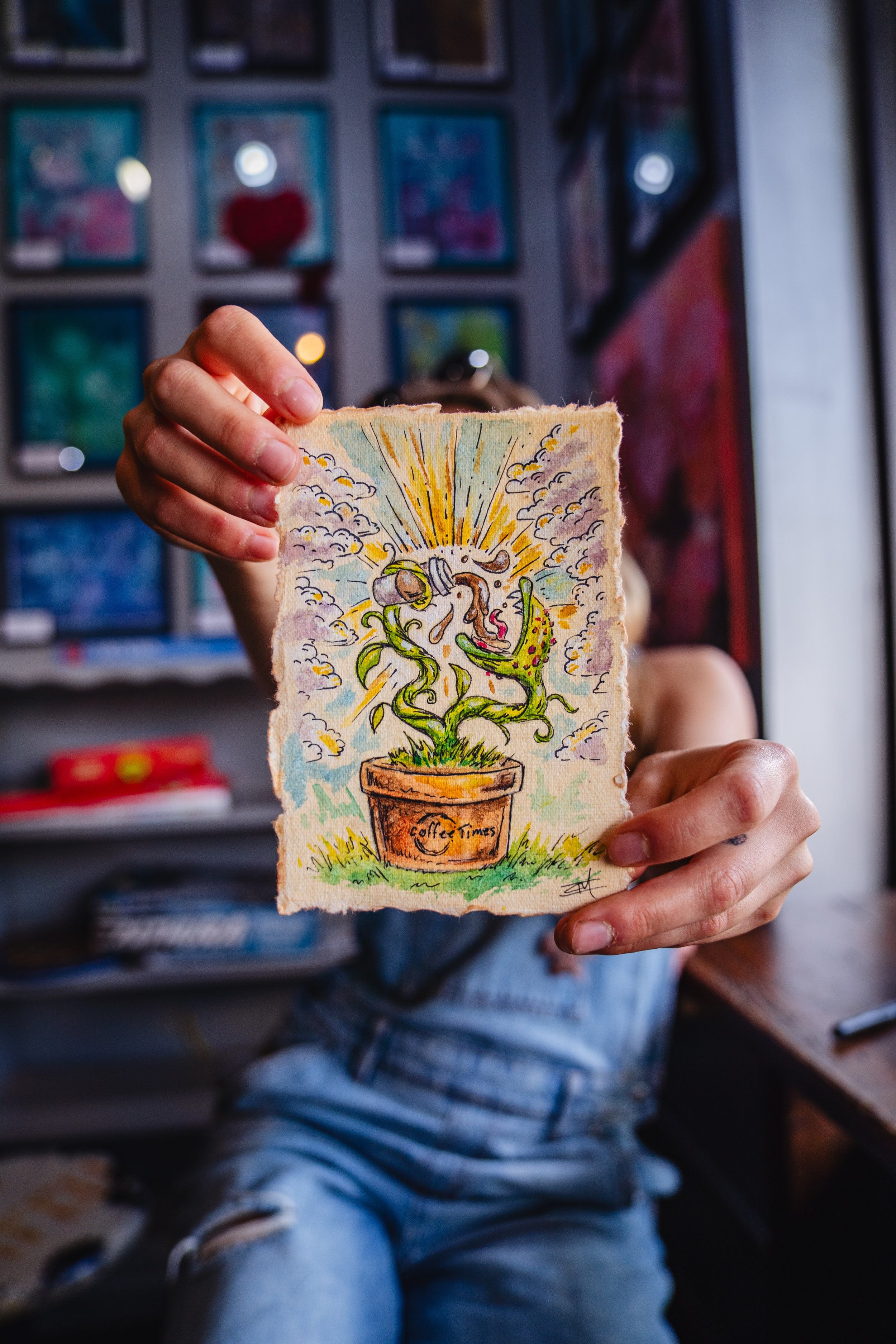 Person holding a colorful drawing of a potted plant labeled "Coffee Times" with whimsical elements and a burst of light, blurred interior background.