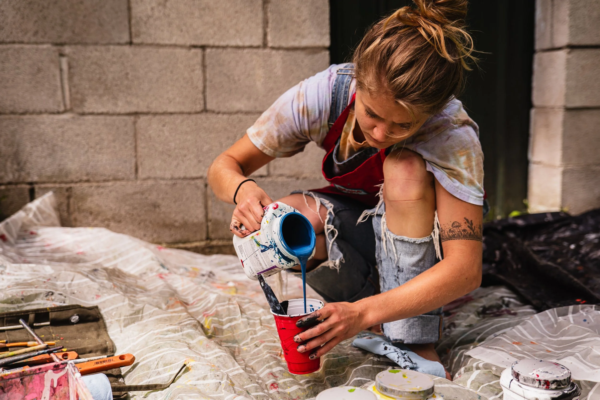 Person pouring blue paint from a can into a red cup on a drop cloth, with additional painting tools nearby.