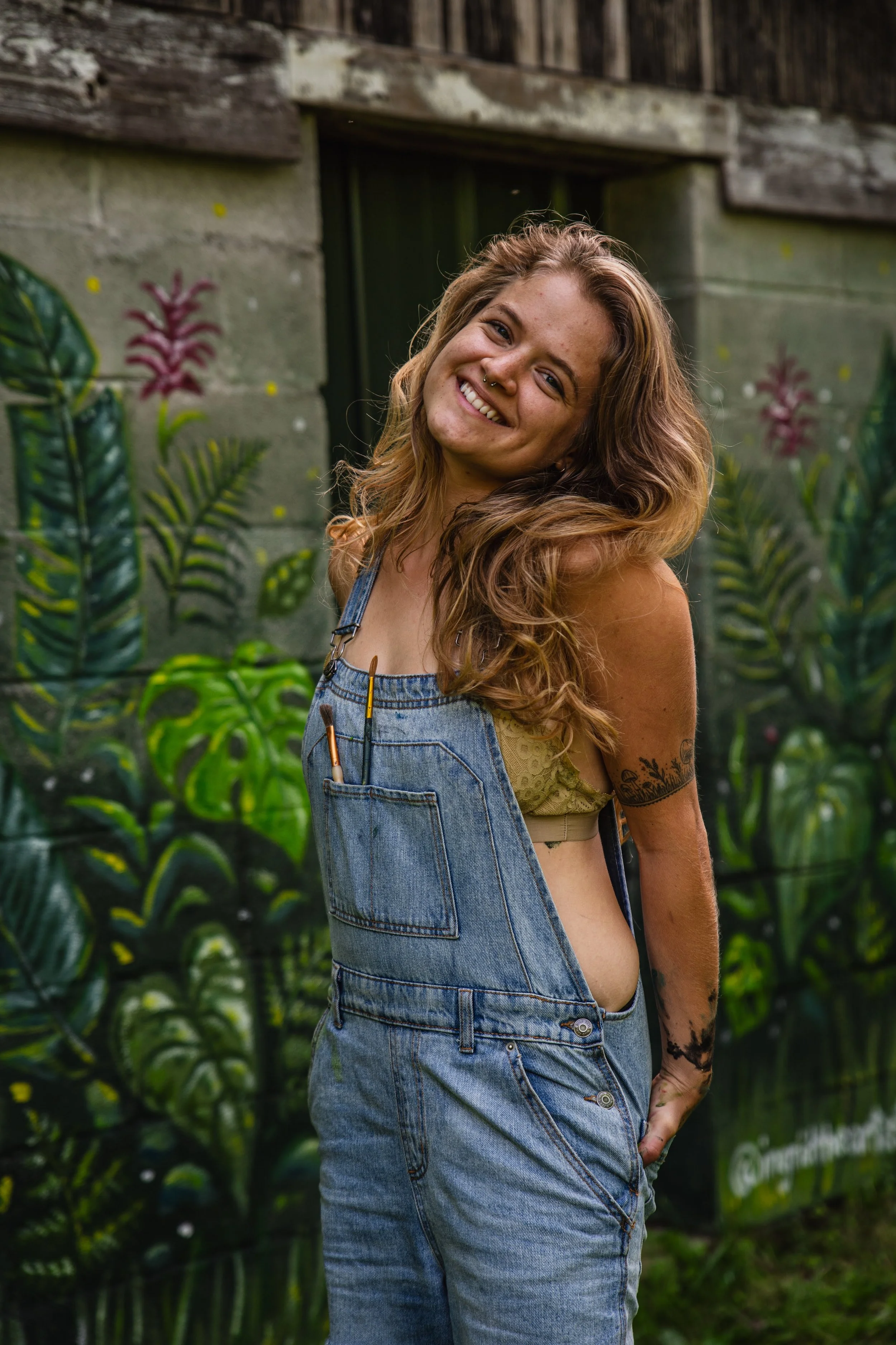 Person smiling in front of a mural with tropical plants, wearing denim overalls and a yellow top.