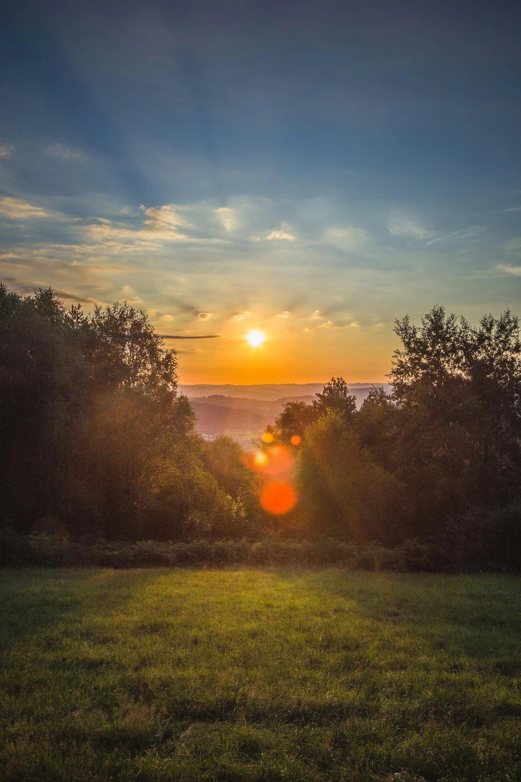 Sunset over a green grassy field with trees on either side and a partly cloudy sky.