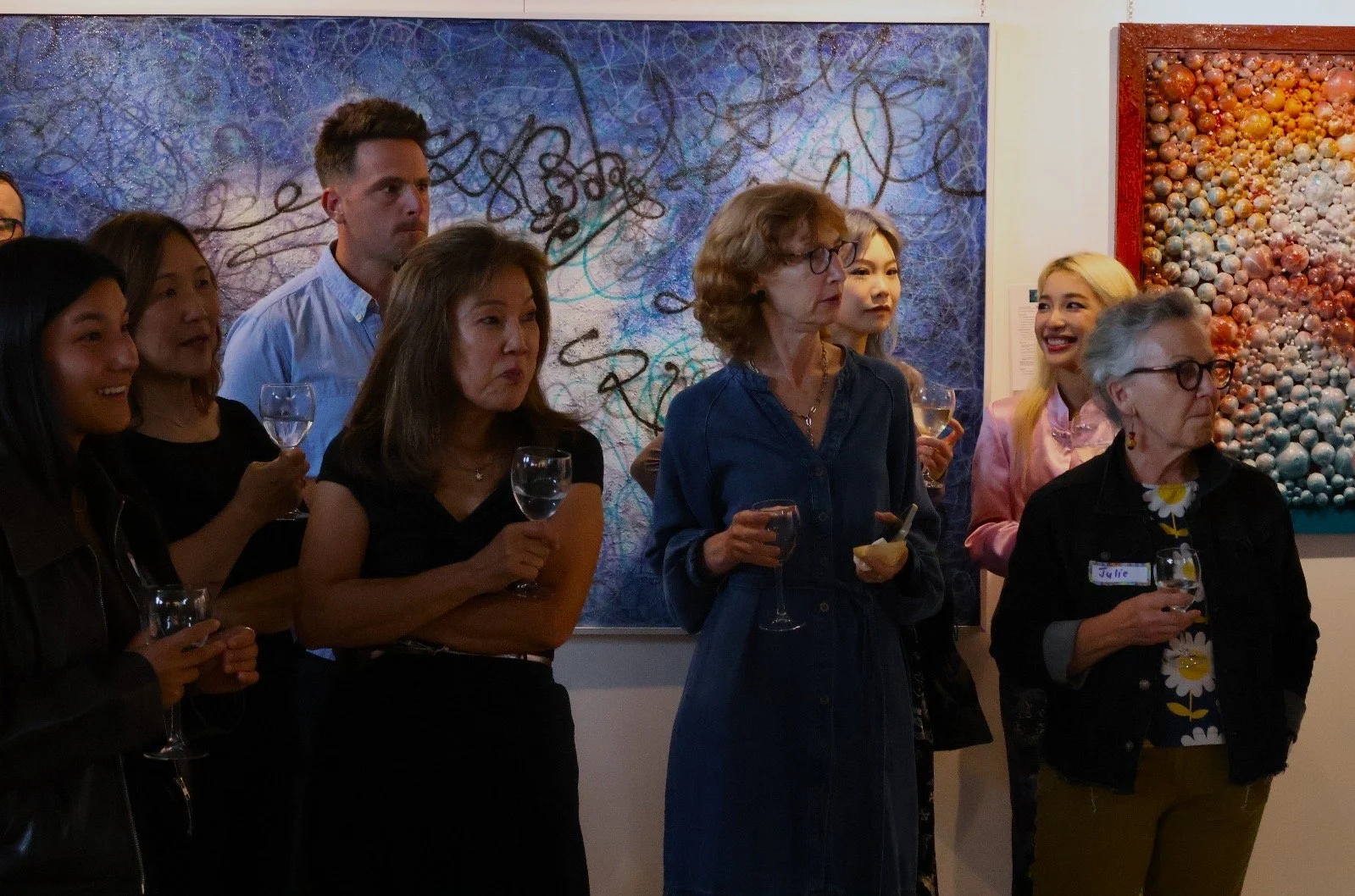 Group of people attending an art gallery event, holding wine glasses, standing in front of abstract paintings on the wall.