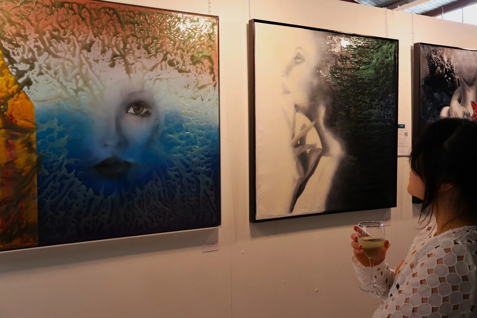 A woman in a white, patterned dress holding a glass of white wine looks at abstract art paintings on a gallery wall.