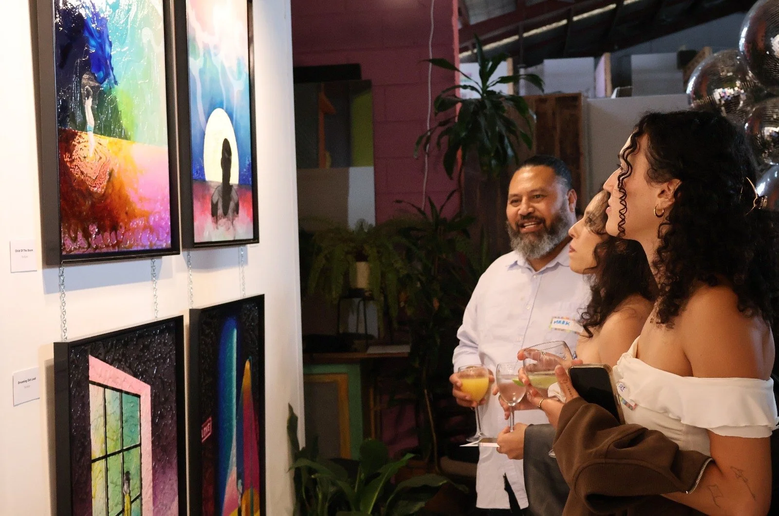 People viewing vibrant, colorful artwork at an art gallery, holding drinks and engaged in conversation.