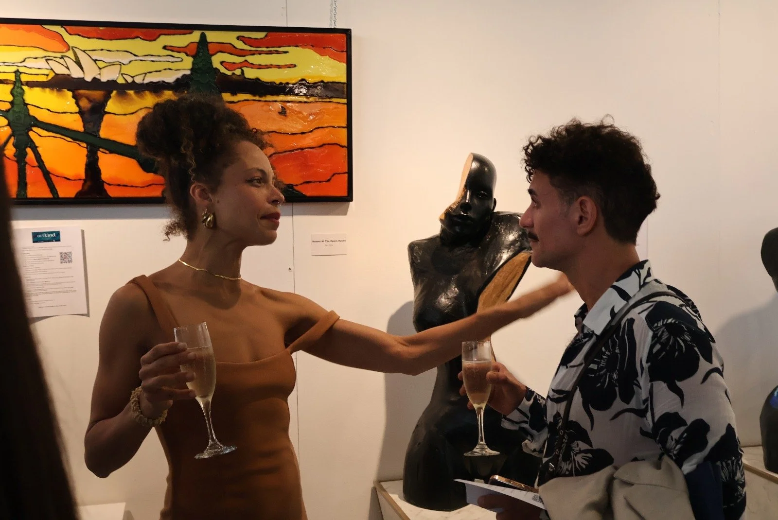 A woman in a brown dress and a man in a black and white floral shirt hold champagne glasses and engage in a conversation at an art gallery. The woman touches the man's face as they look at each other, with artwork and sculptures visible in the backgr