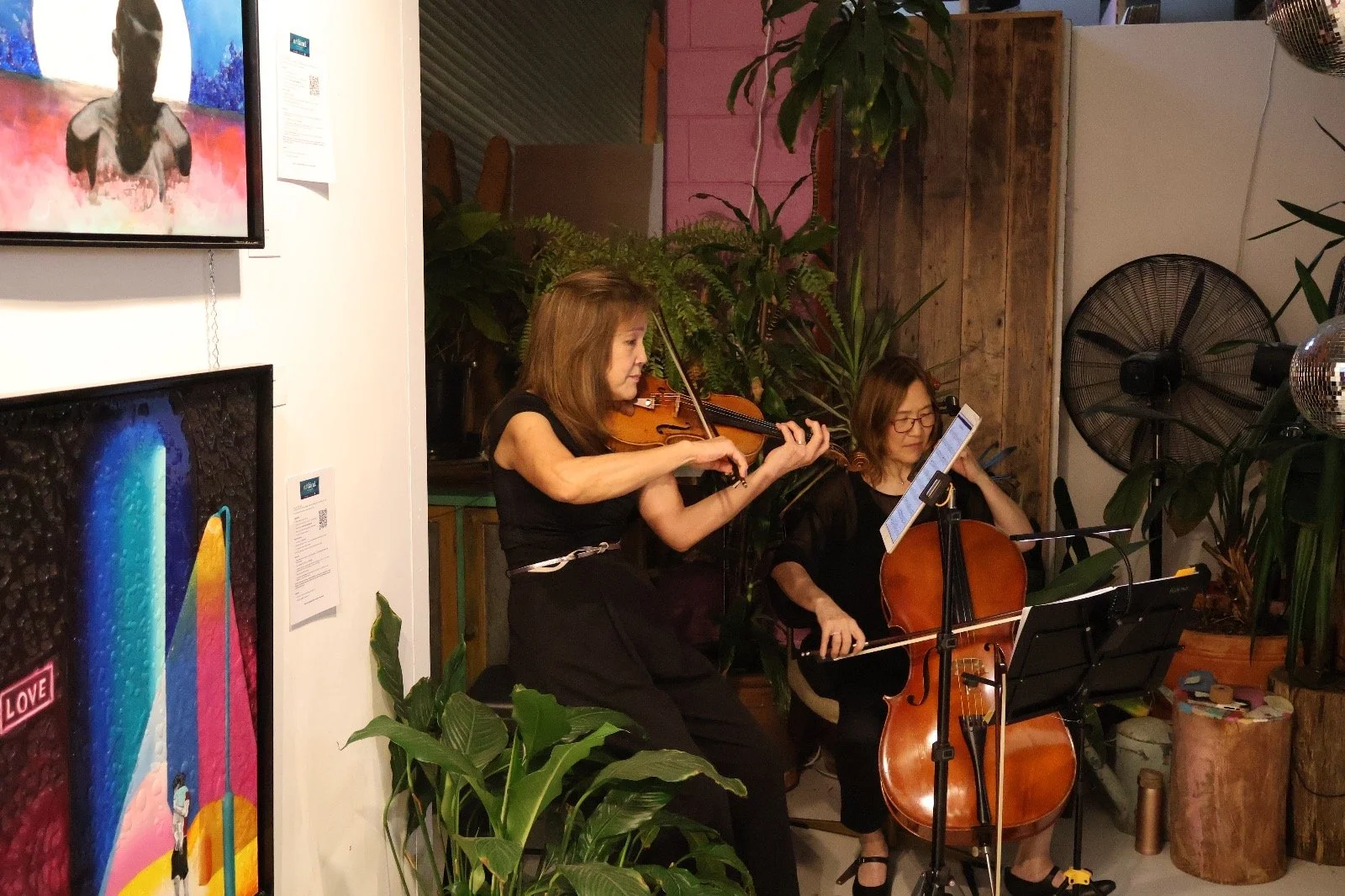 Two women play string instruments, a violin and a cello, in an indoor setting surrounded by green plants and art on the walls.