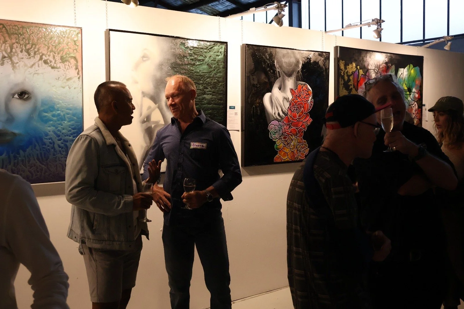 People chatting at an art exhibit with several abstract portrait paintings displayed on the wall.