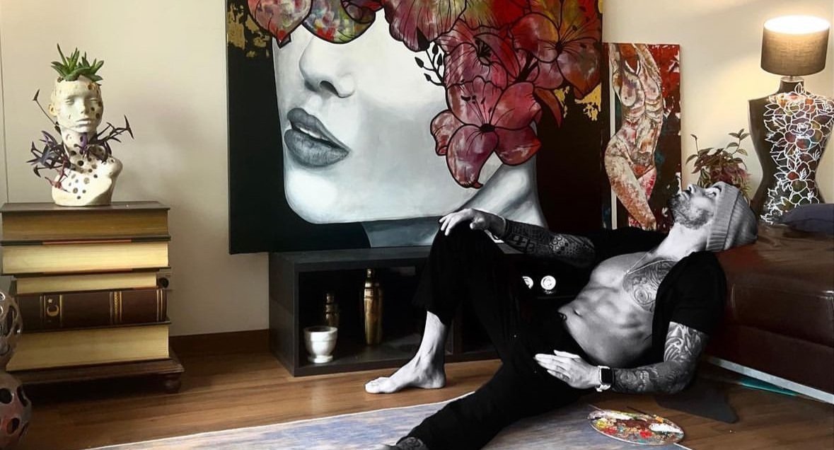 Daniele D'Aluisio, reclining on a couch in a living room with art deco and floral decor, including a large portrait of a woman's face with floral hair and a bust with a plant on top.
