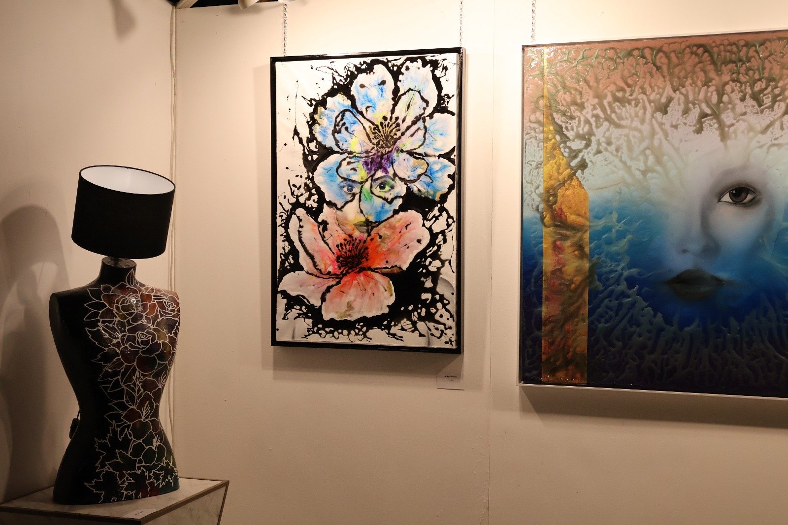 An art gallery with two colorful paintings on the wall, a black table lamp with a female mannequin base decorated with floral outlines, and the paintings feature abstract floral and face designs.