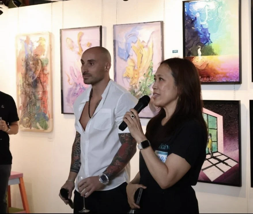 A woman speaking into a microphone at an art gallery, with artwork hanging behind her. A man standing next to her, holding a glass of wine. Other people partially visible in the background.
