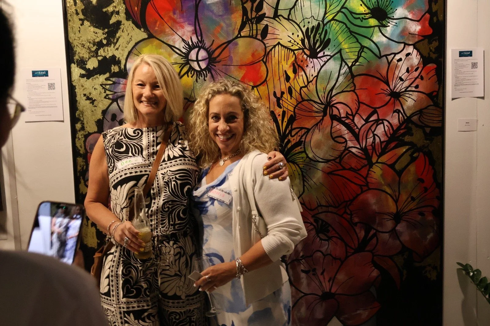 Two women smiling and posing for a photo at an art gallery, standing in front of a colorful floral painting. One woman has blonde hair and is wearing a black and white patterned dress, holding a drink; the other has curly blonde hair, wearing a white