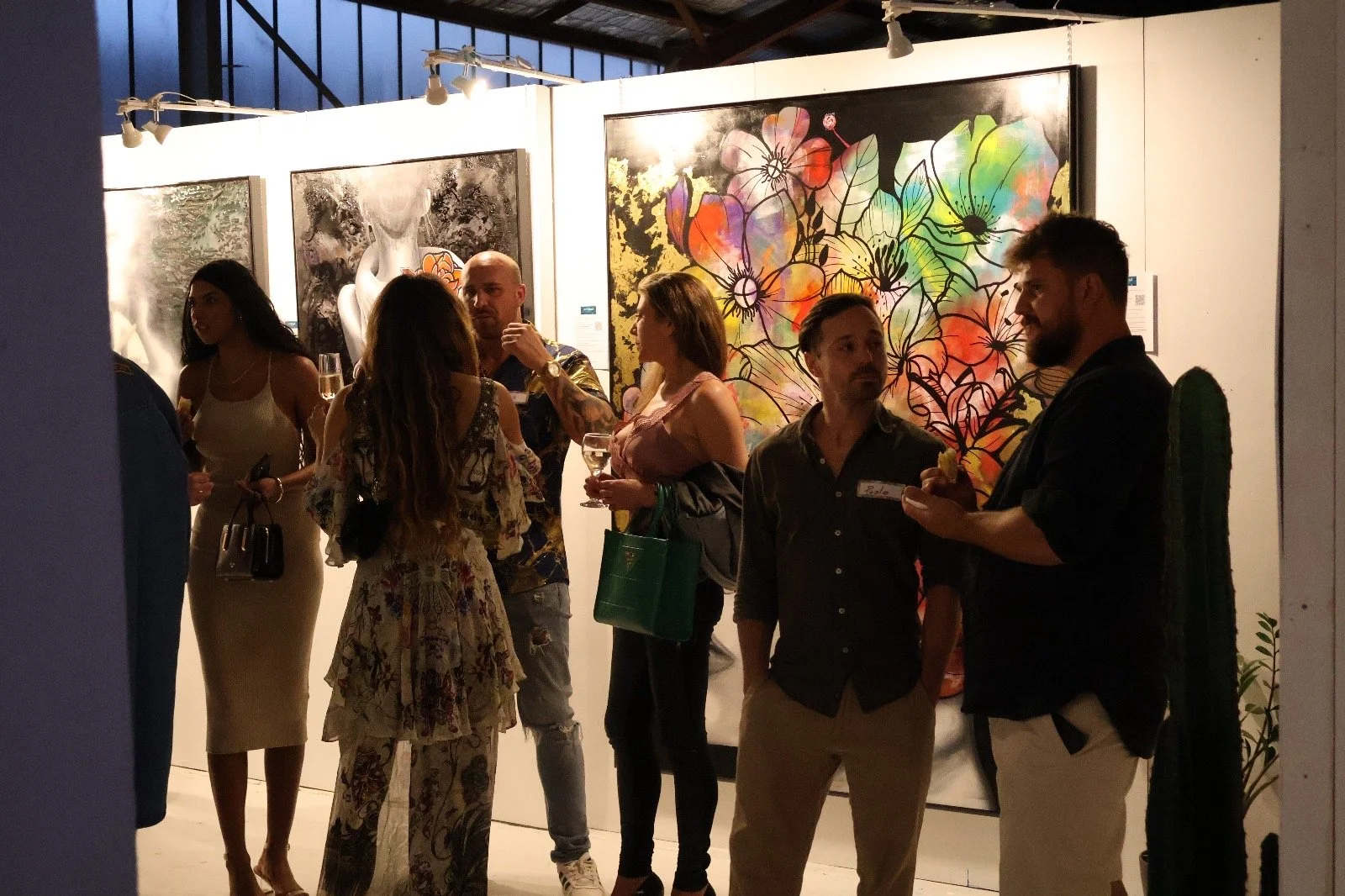 People attending an art gallery exhibition, engaging in conversation near colorful paintings on display.