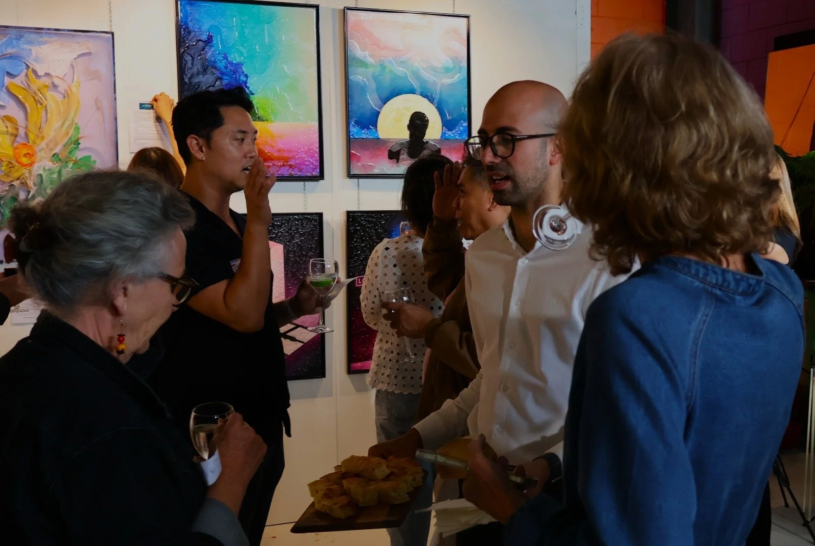 People socializing at an art gallery, with colorful abstract paintings on the wall, holding drinks and snacks.