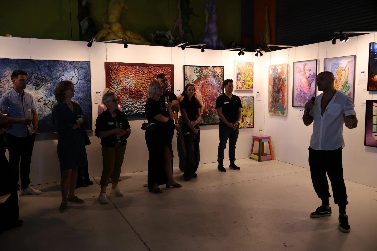 Gallery opening with Daniele D'Aluisio giving a speech, audience holding drinks, and colorful abstract paintings on white walls.