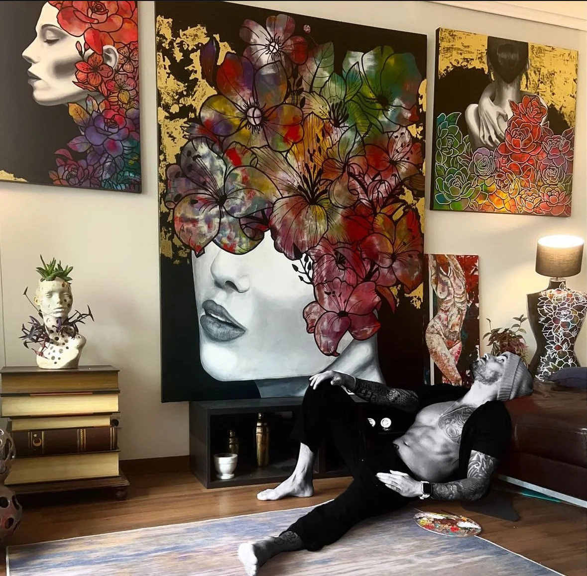 A man with tattoos and wearing a beanie hat reclines on the floor in a room filled with colorful art. Behind him is a large black and white painting of a woman's face with flowers covering her eyes. The room features additional floral artwork, a bust with plants on top, a lamp, and decorative objects.