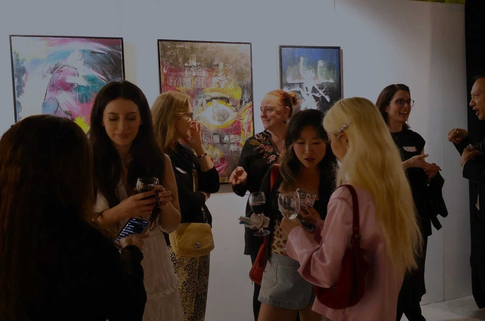 Group of women and one man at an art gallery, some holding wine glasses, engaging in conversation and looking at artworks.