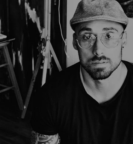 Black and white photo of artist Daniele D'Aluisio wearing glasses and a cap, with artistic equipment visible in the background.