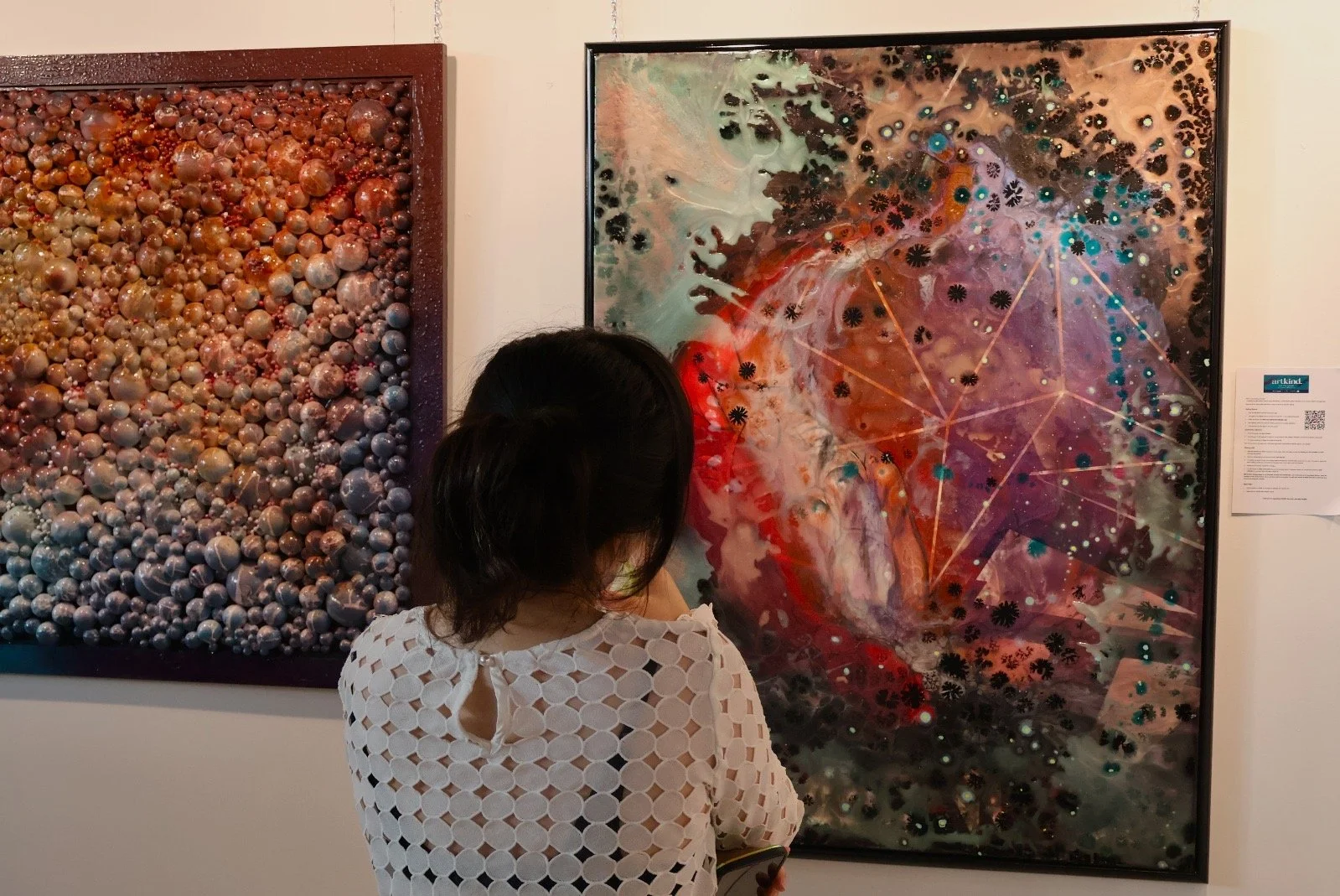 A woman with short dark hair and a white patterned top is standing in front of abstract paintings in a gallery, looking at a vibrant, multicolored piece with geometric lines and floral motifs.