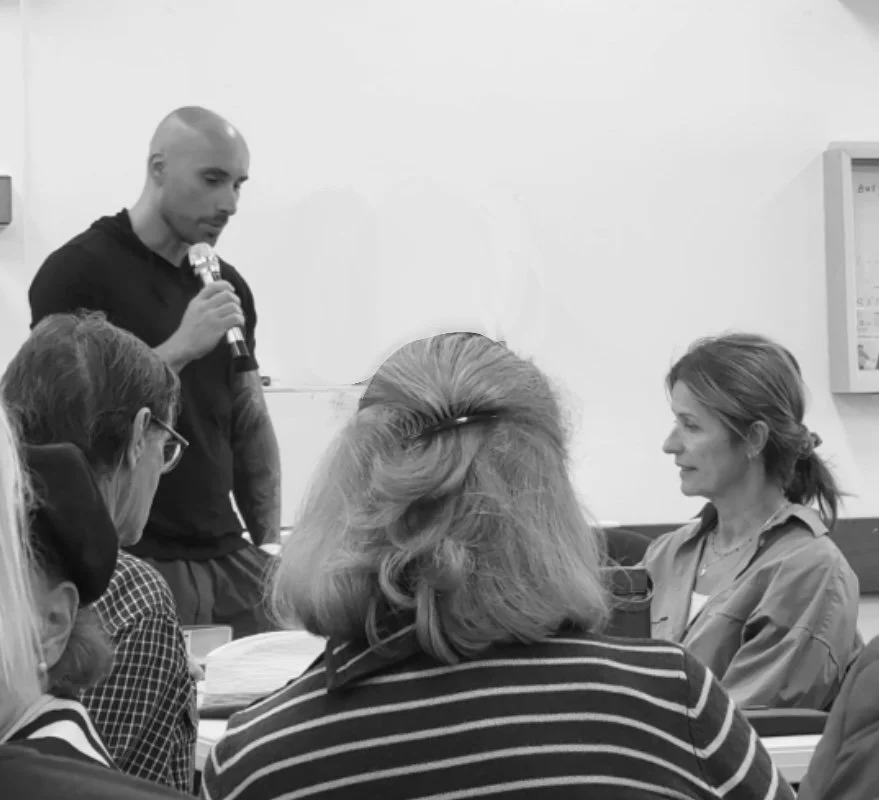 Artist Daniele D'Aluisio with a microphone speaking to a seated woman in a room with several other people.