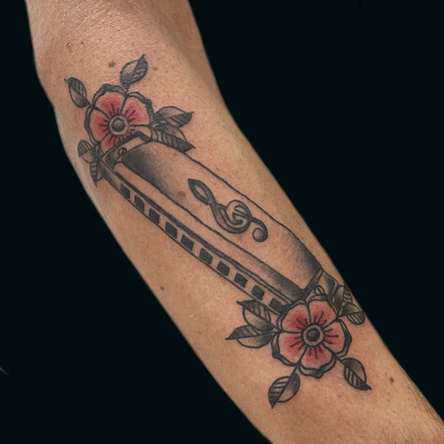 Tattoo of a guitar in black and gray with pink flowers surrounding it on a person's arm.