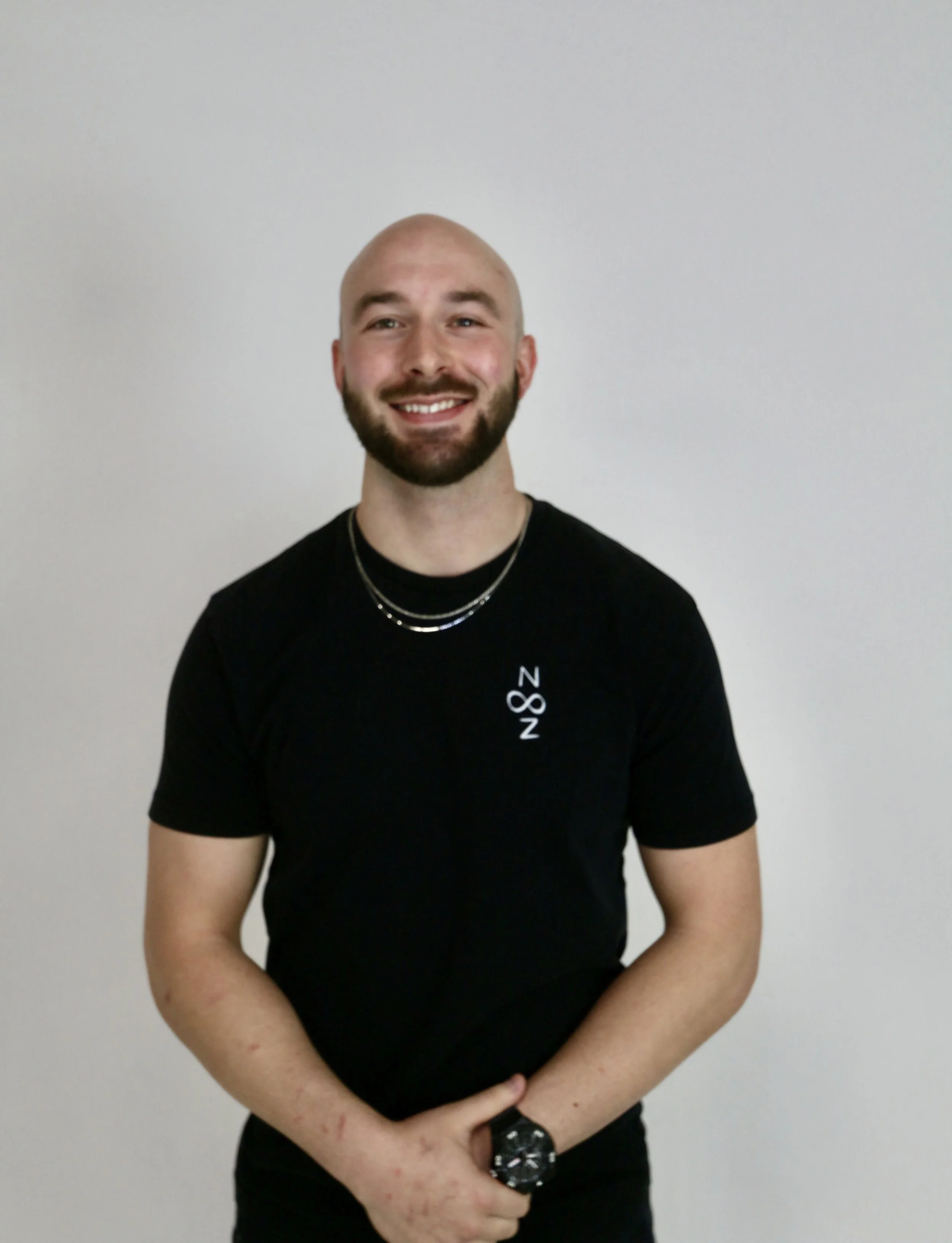 A smiling man with a beard and shaved head wearing a black t-shirt with white text, two silver necklaces, and a watch standing against a plain wall.