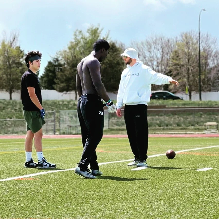 Three men standing on a football field, two of them in a huddle and one observing, with a football on the ground nearby and trees in the background.