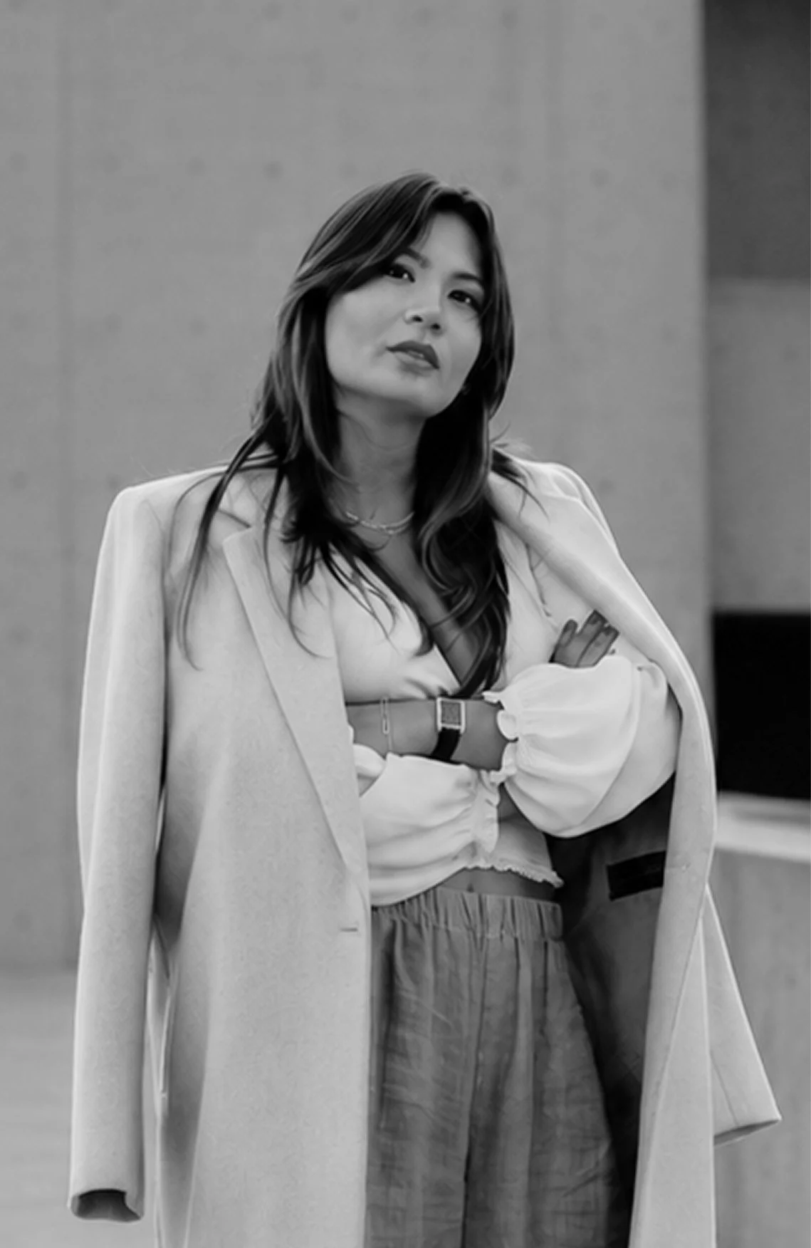 A woman with long dark hair wearing a light-colored blazer and patterned pants, standing against a plain background.