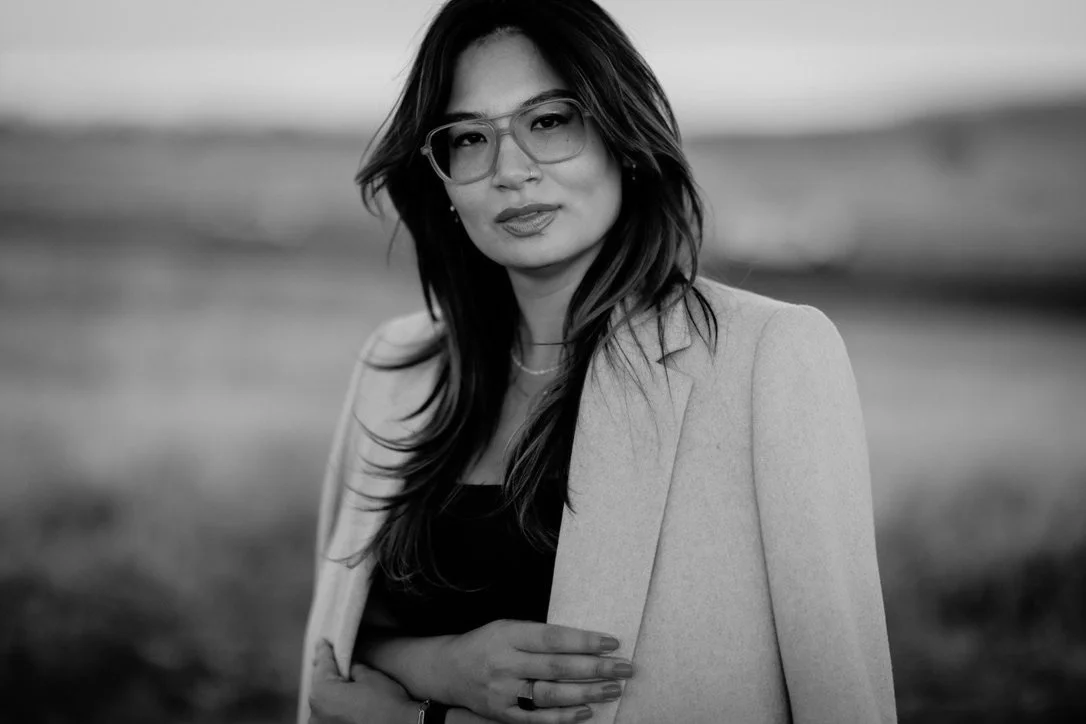 Black and white photo of a woman wearing glasses and a light-colored blazer, standing outdoors with a blurred background.