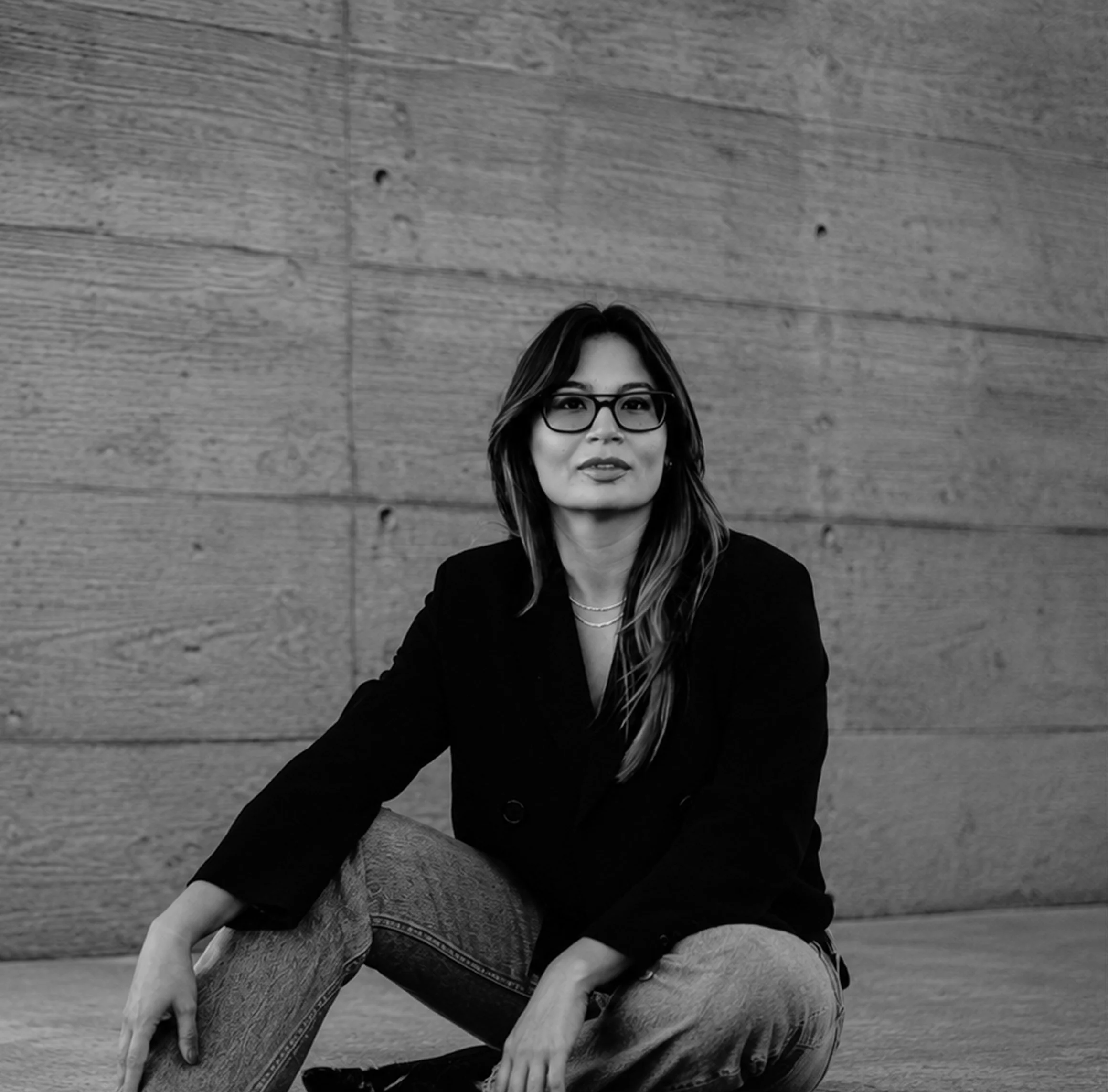 A woman with glasses sitting on a wooden floor, wearing a black blazer and patterned scarf, smiling at the camera.
