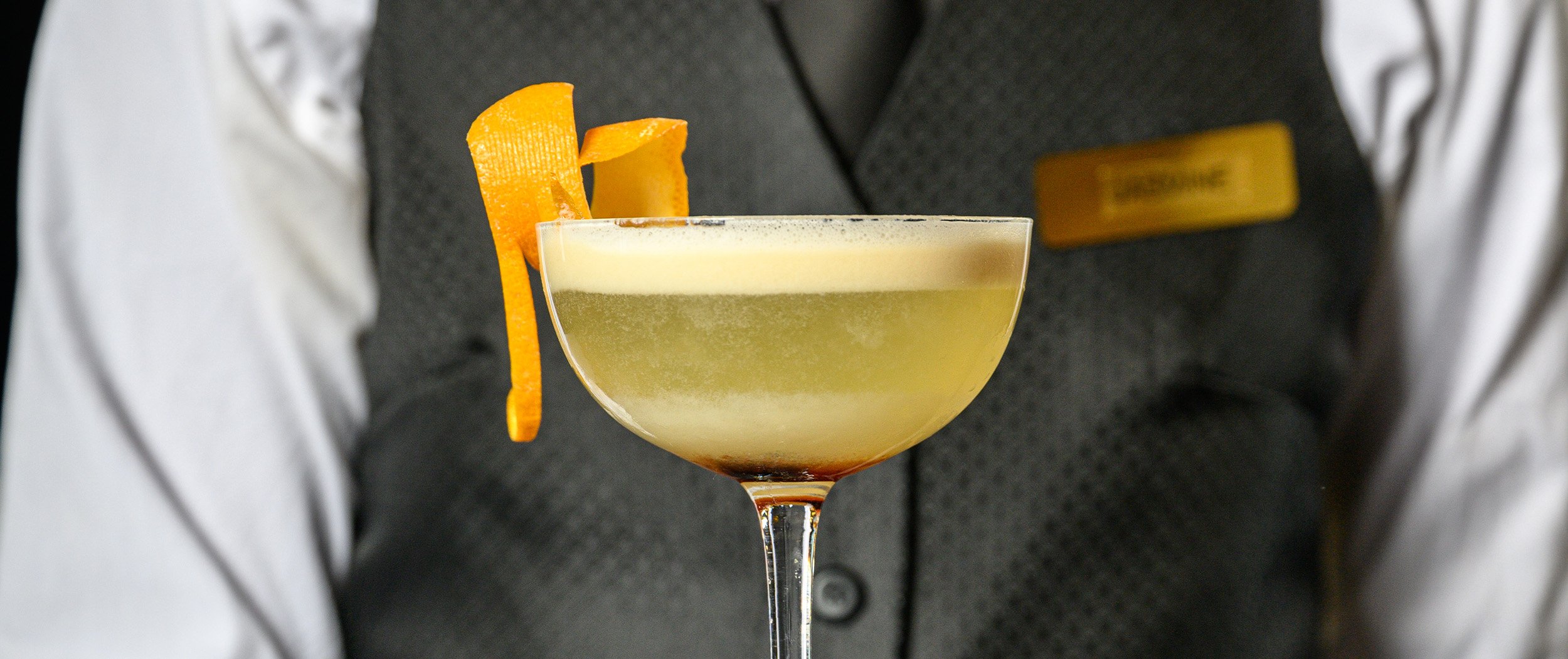A cocktail garnished with an orange peel, served in a coupe glass held by a person wearing a vest and white shirt.