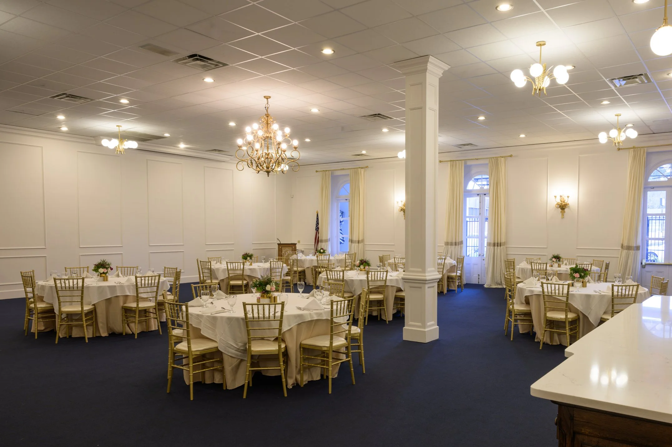 A banquet hall with round tables covered in white tablecloths, set with glassware, silverware, and napkins. Chandeliers and pendant lights illuminate the room, which features dark chairs and decorative wall mirrors.