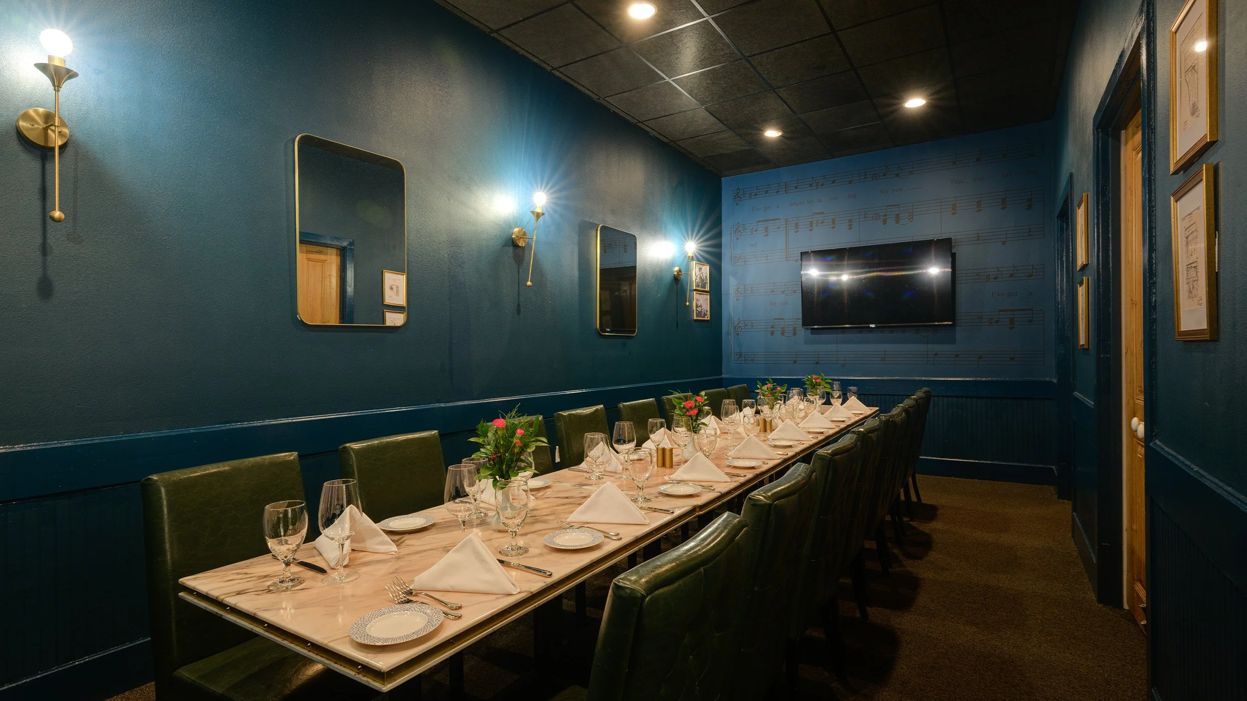 Elegant dining room with a long table set for a formal meal. The room has a dark blue wall with decorative sconces, mirrors, and framed art. The table is adorned with white napkins, glassware, and floral centerpieces. A flat-screen TV is mounted on the wall.