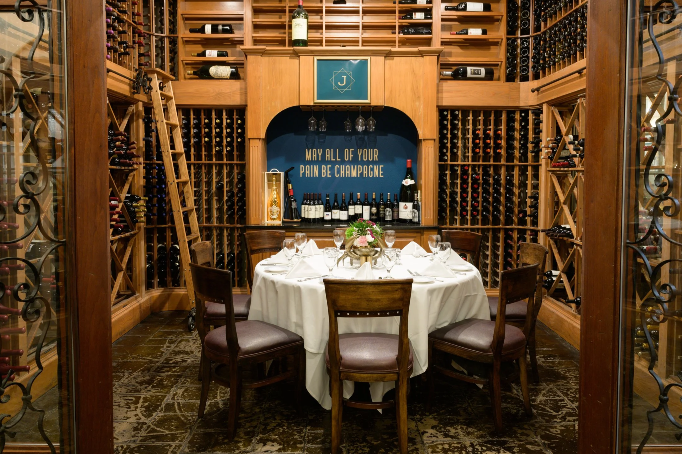 A wine cellar with wooden racks filled with wine bottles surrounding a round dining table set with white tablecloth, glassware, and cutlery. A ladder is leaning against the racks, and a sign on the wall reads "May all of your pain be Champagne."
