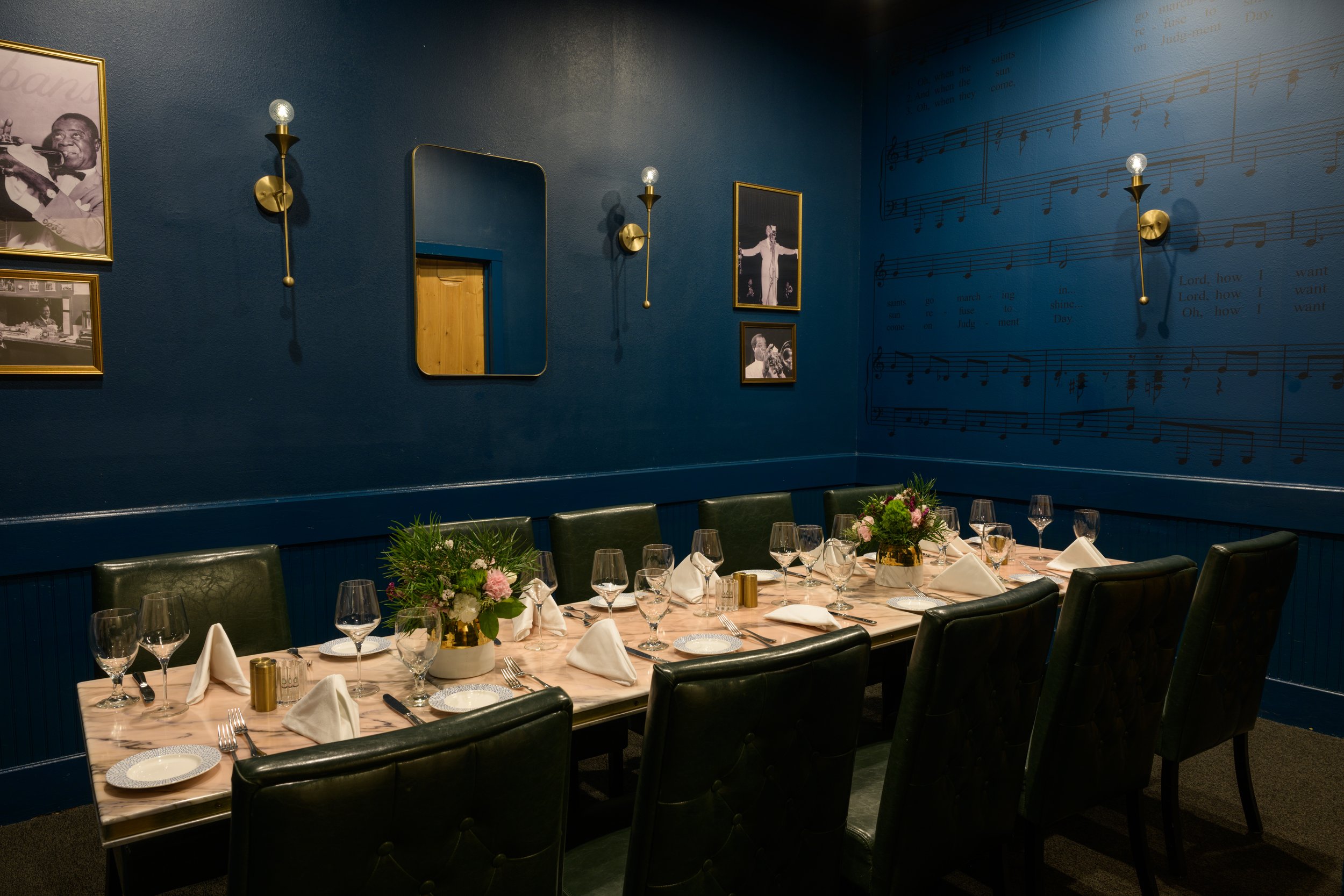 A dimly lit, elegantly set dining room with a long table and eight chairs. The walls are deep blue, adorned with framed photographs and sheet music. There are sconces providing soft lighting, and the table is prepared with white napkins, glassware, plates, and small centerpieces with red flowers.